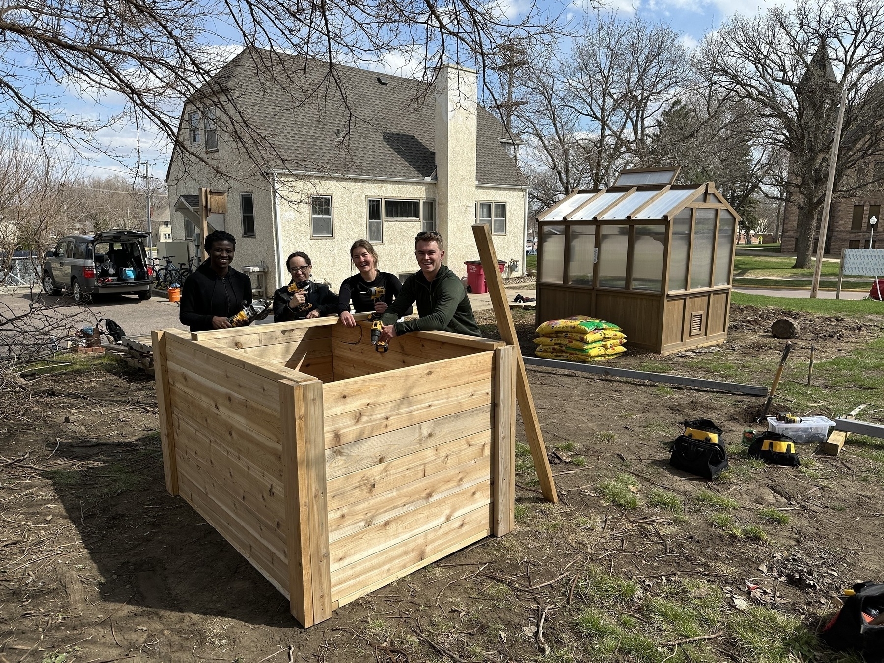 Auto-generated description: A group of people are constructing a wooden structure outdoors near a house and greenhouse.