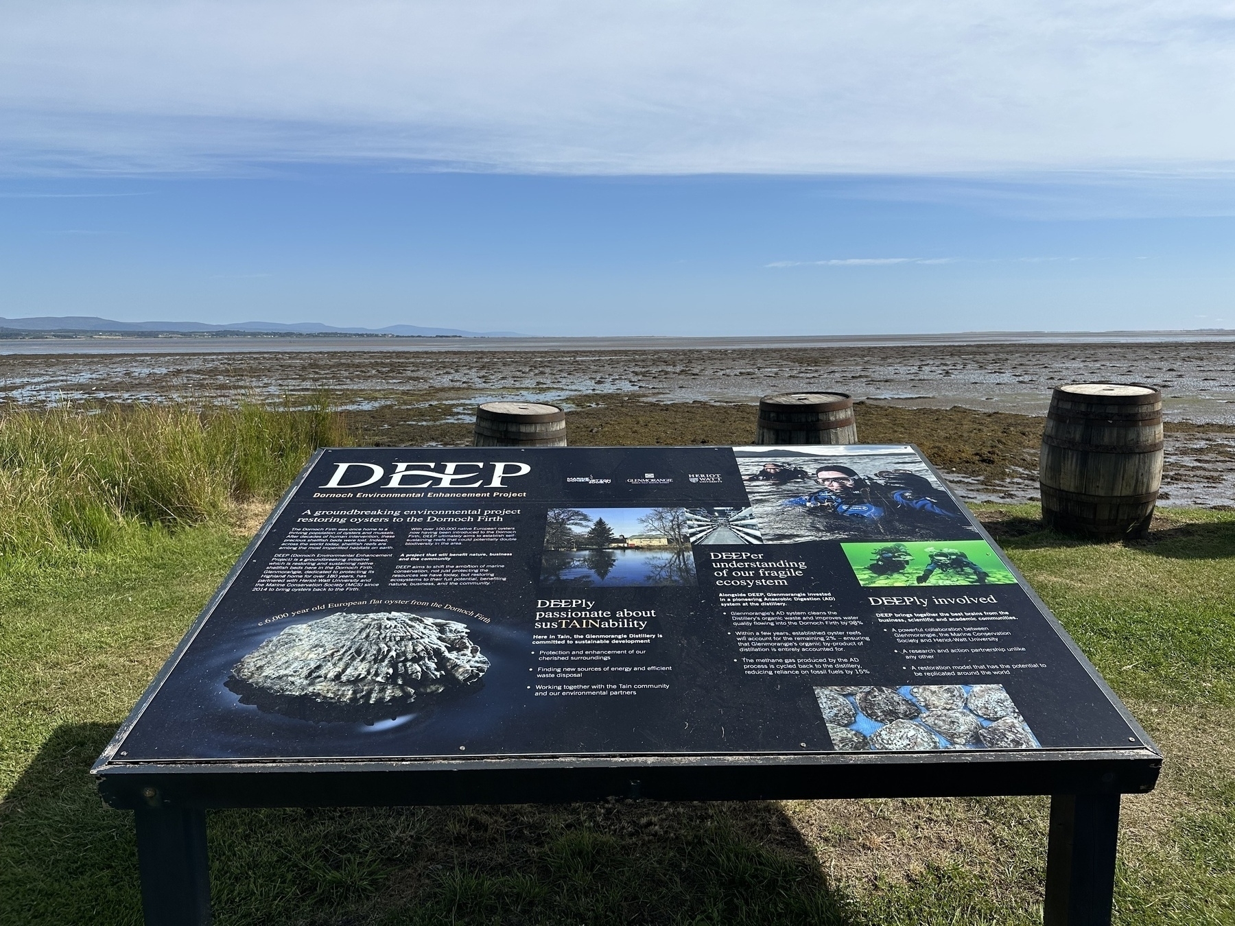 An informational sign about marine conservation is situated by a coastal area with barrels and a view of the sea in the background.