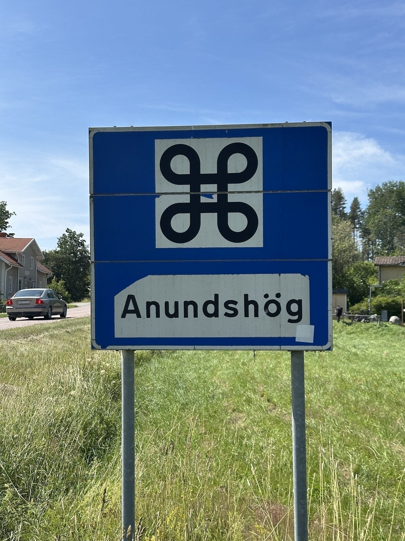 A road sign displays the name "Anundshög" beneath a symbol resembling a looped square or St. John's Arms on a blue background, set in a grassy area with houses and a car in the distance.