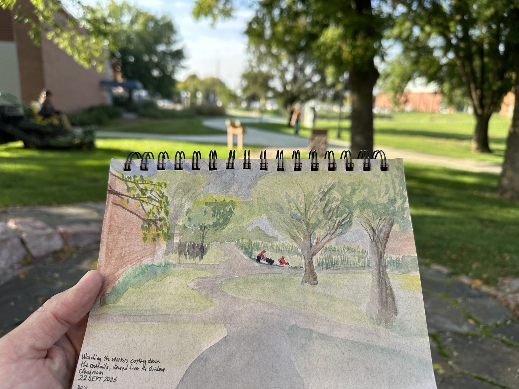 A hand holds a sketchpad displaying a watercolor painting of a park scene that closely matches the actual park in the background.