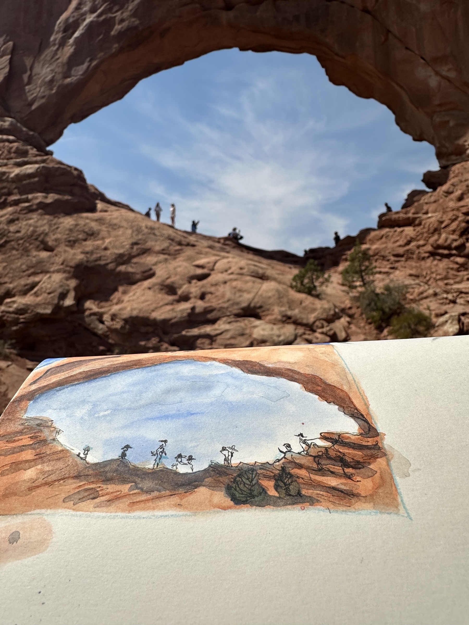 A watercolor painting of a natural rock arch is being held against the backdrop of the same arch, with people visible on it.