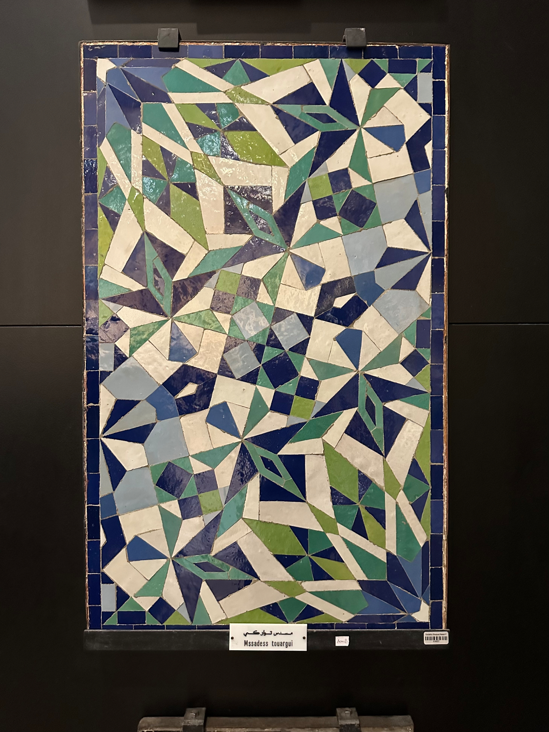 A mosaic artwork features an abstract geometric pattern in shades of blue, green, and white, displayed on a dark background.