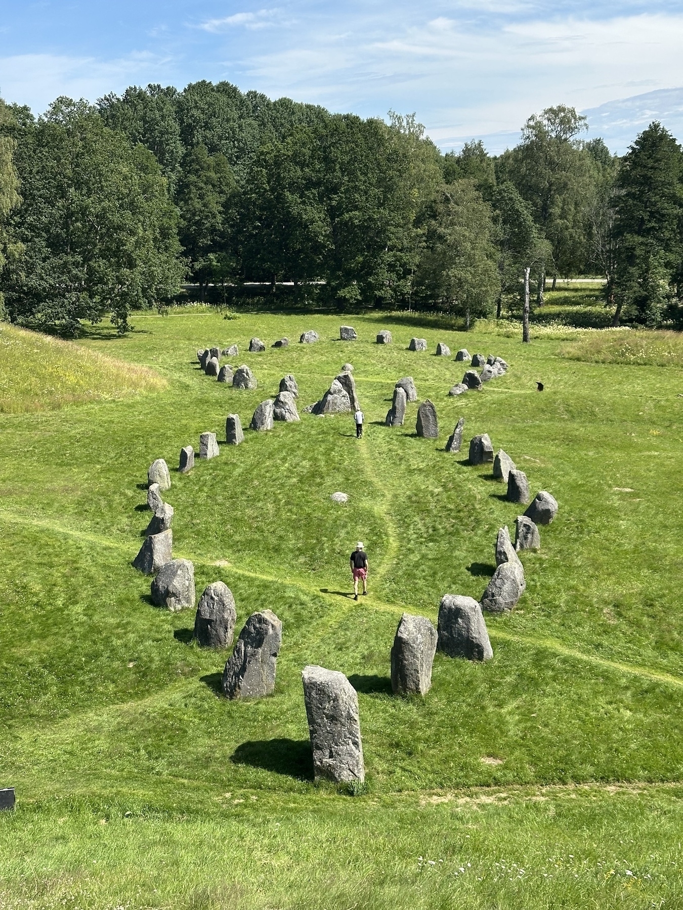 A person walks through a grassy field surrounded by a large, elongated stone circle formation amidst a backdrop of trees.