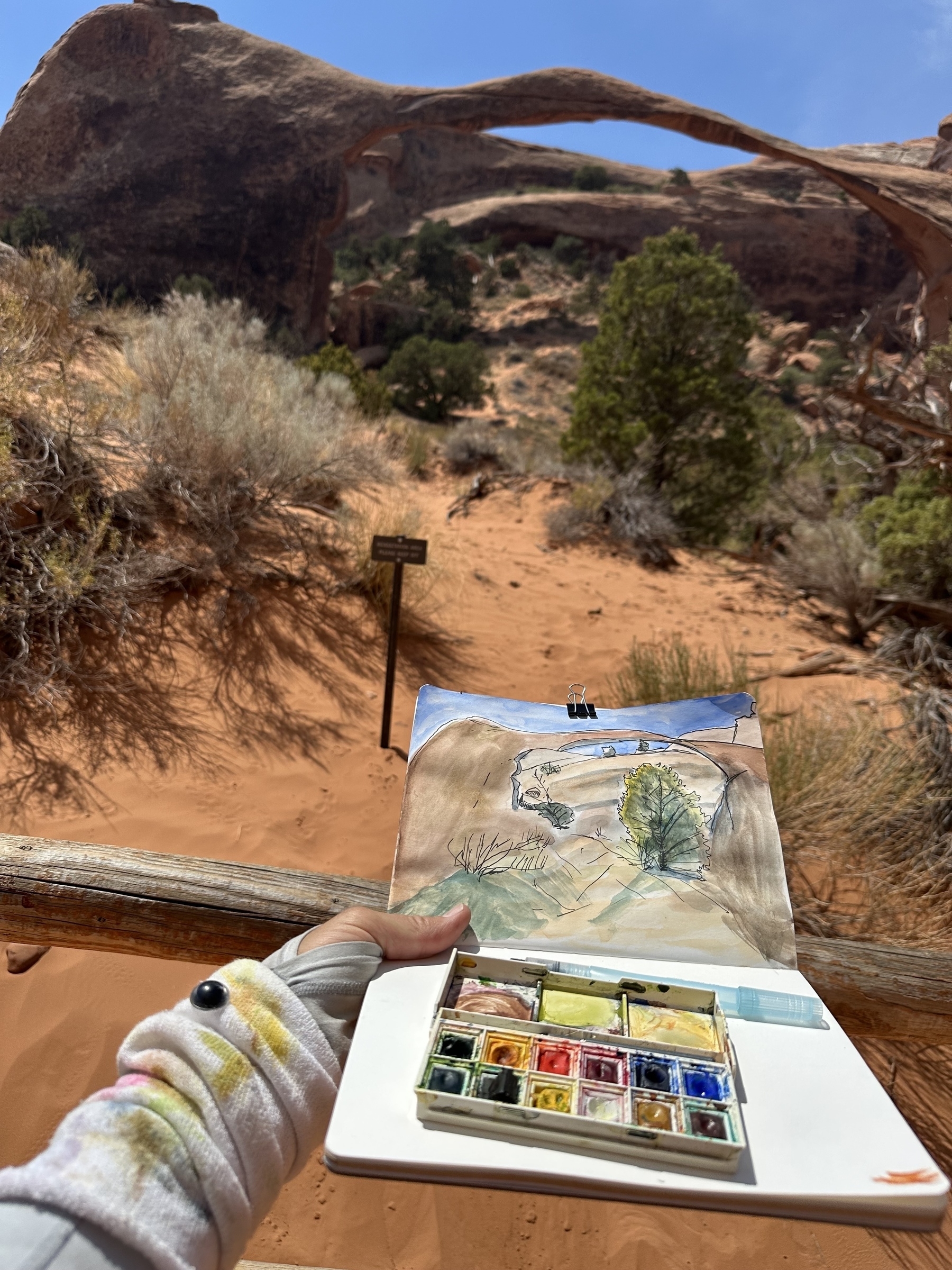 A person is painting a desert landscape with natural arches using watercolors, while holding the painting against the backdrop of the scene being painted.