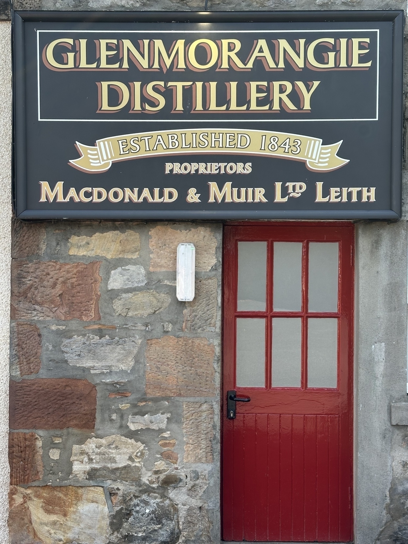 A red door is set in a stone wall beneath a sign for Glenmorangie Distillery.