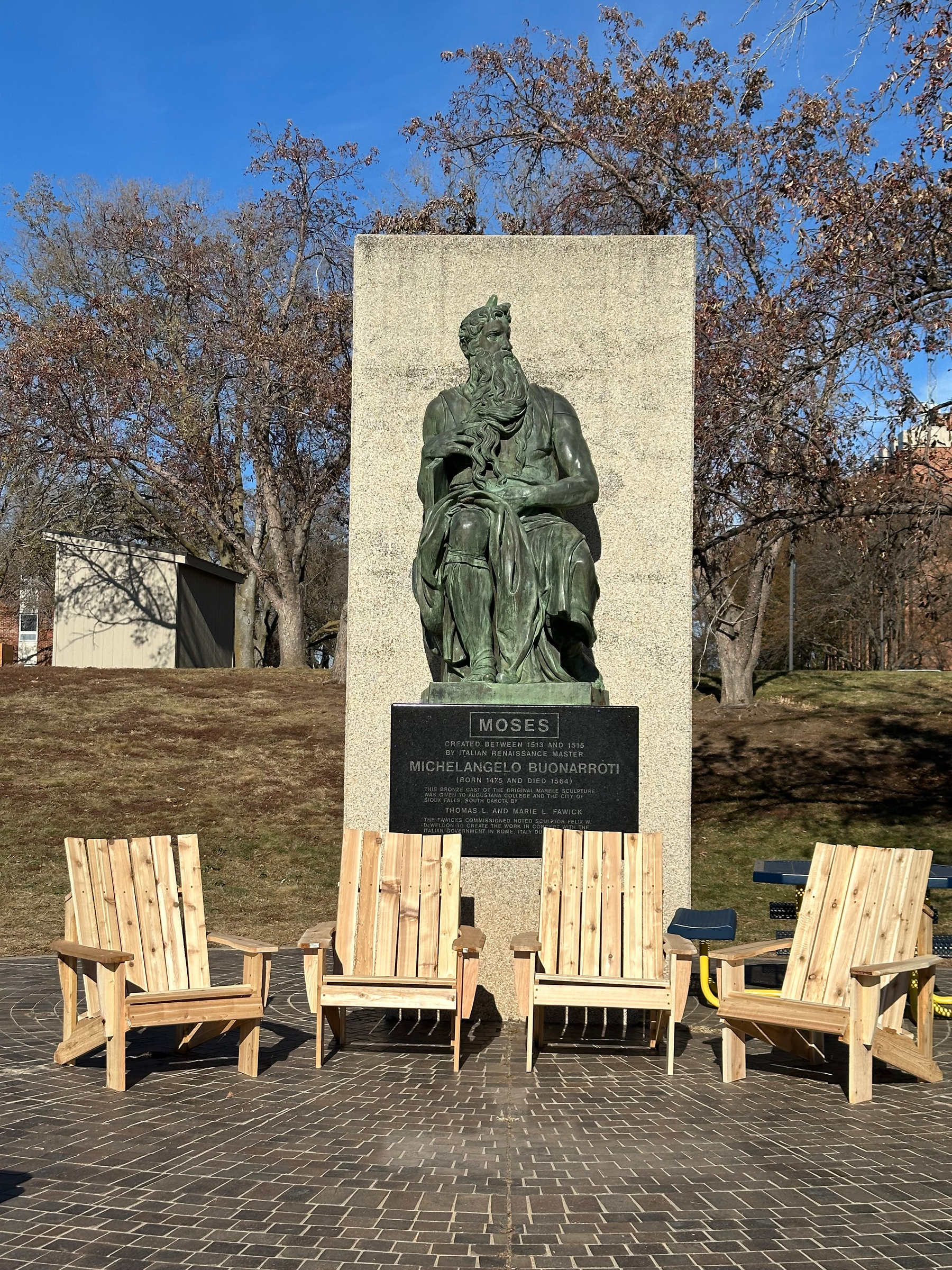 A statue of a seated figure, identified as Michelangelo's Moses, is displayed on a stone pedestal, surrounded by wooden chairs on a brick patio.