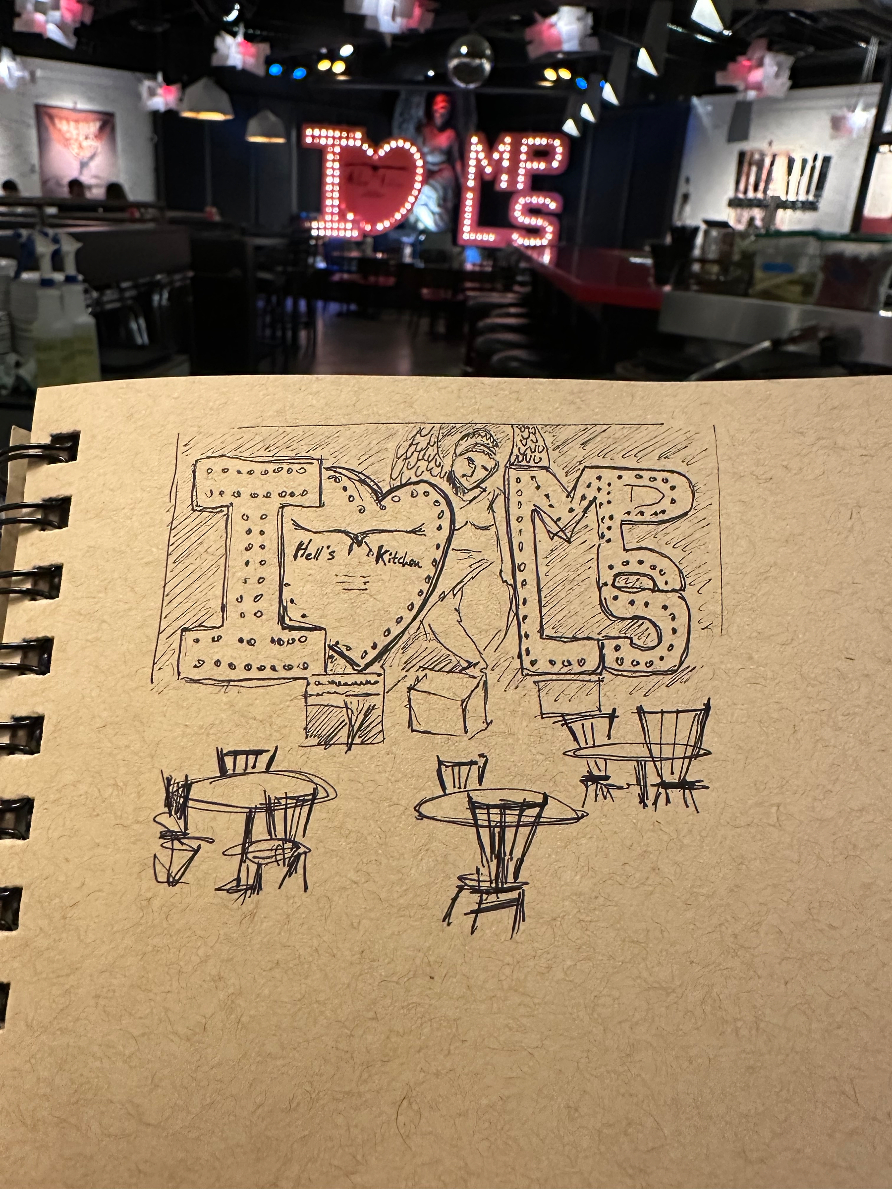 A sketch on a notepad captures a cafe scene with tables, chairs, and a large I ♥ MPLS sign, closely mirroring the illuminated and decorated environment visible in the background.