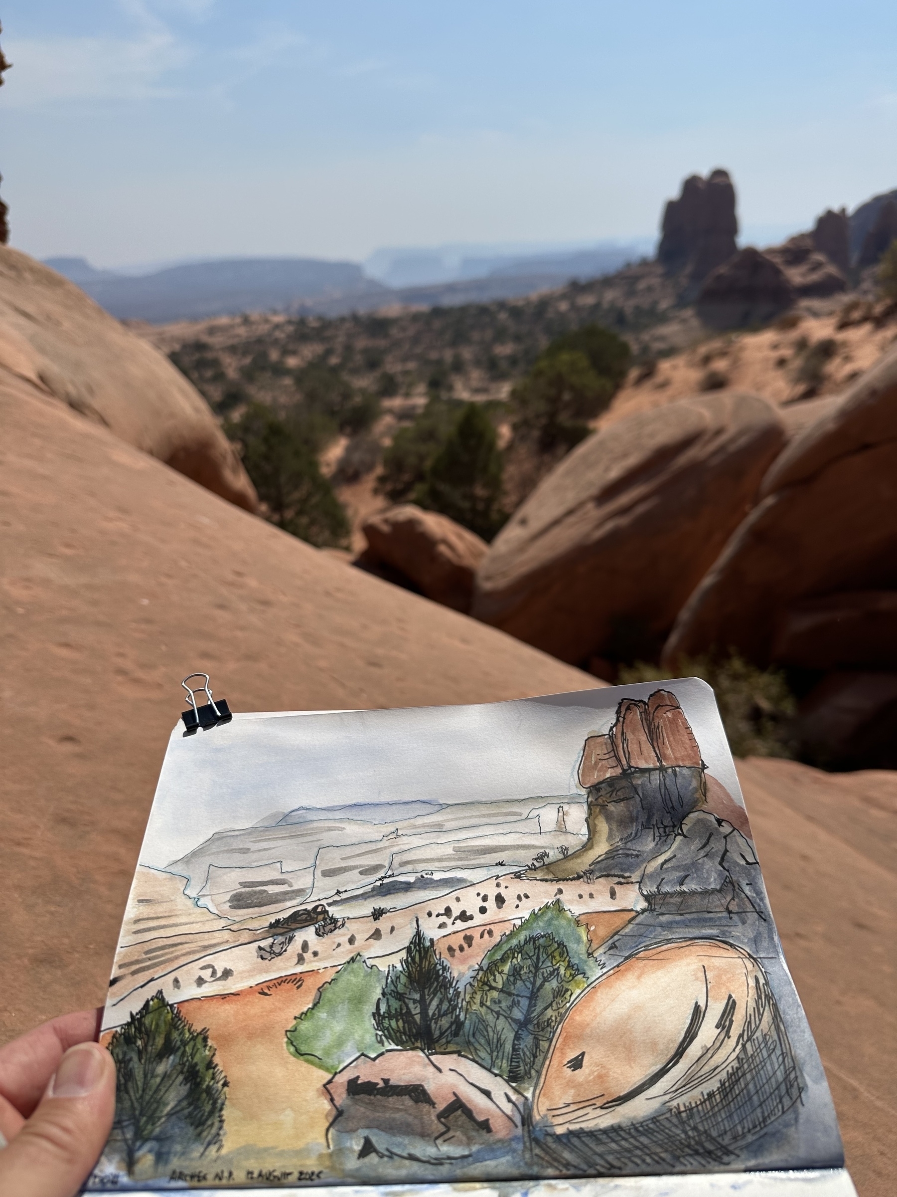 A person holds a sketch of a desert landscape with red rock formations, aligning it with the real view in the background.
