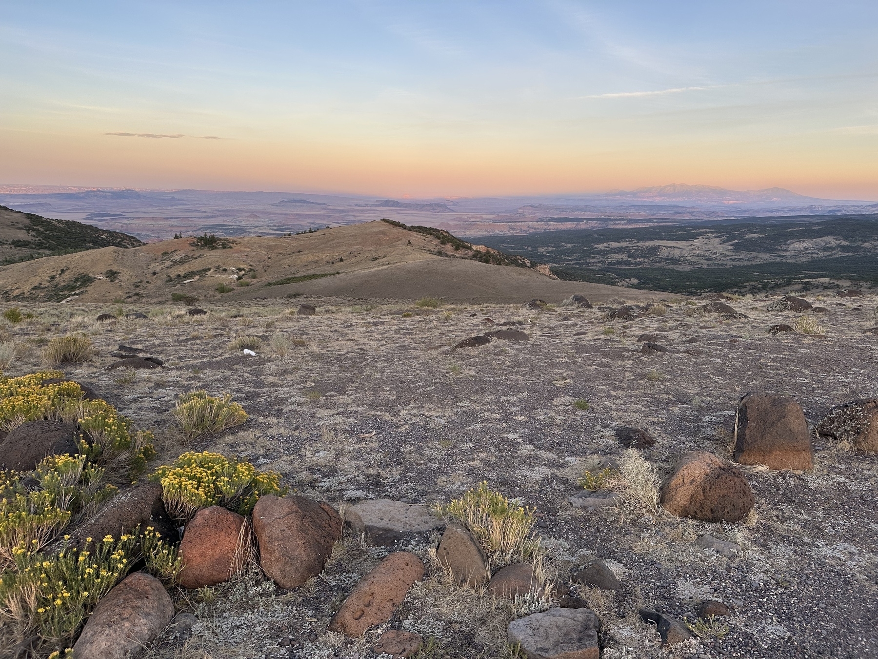 Dry mountain vista at sunset with rainbow colors across the sky and small rocks and vegetation at the foreground