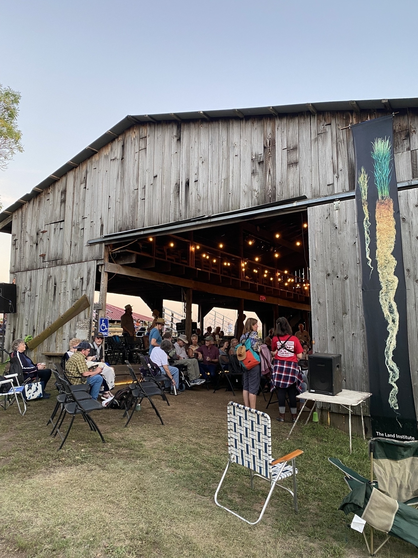 A group of people is gathered inside and around a rustic wooden barn decorated with string lights, while others are seated on chairs outside.