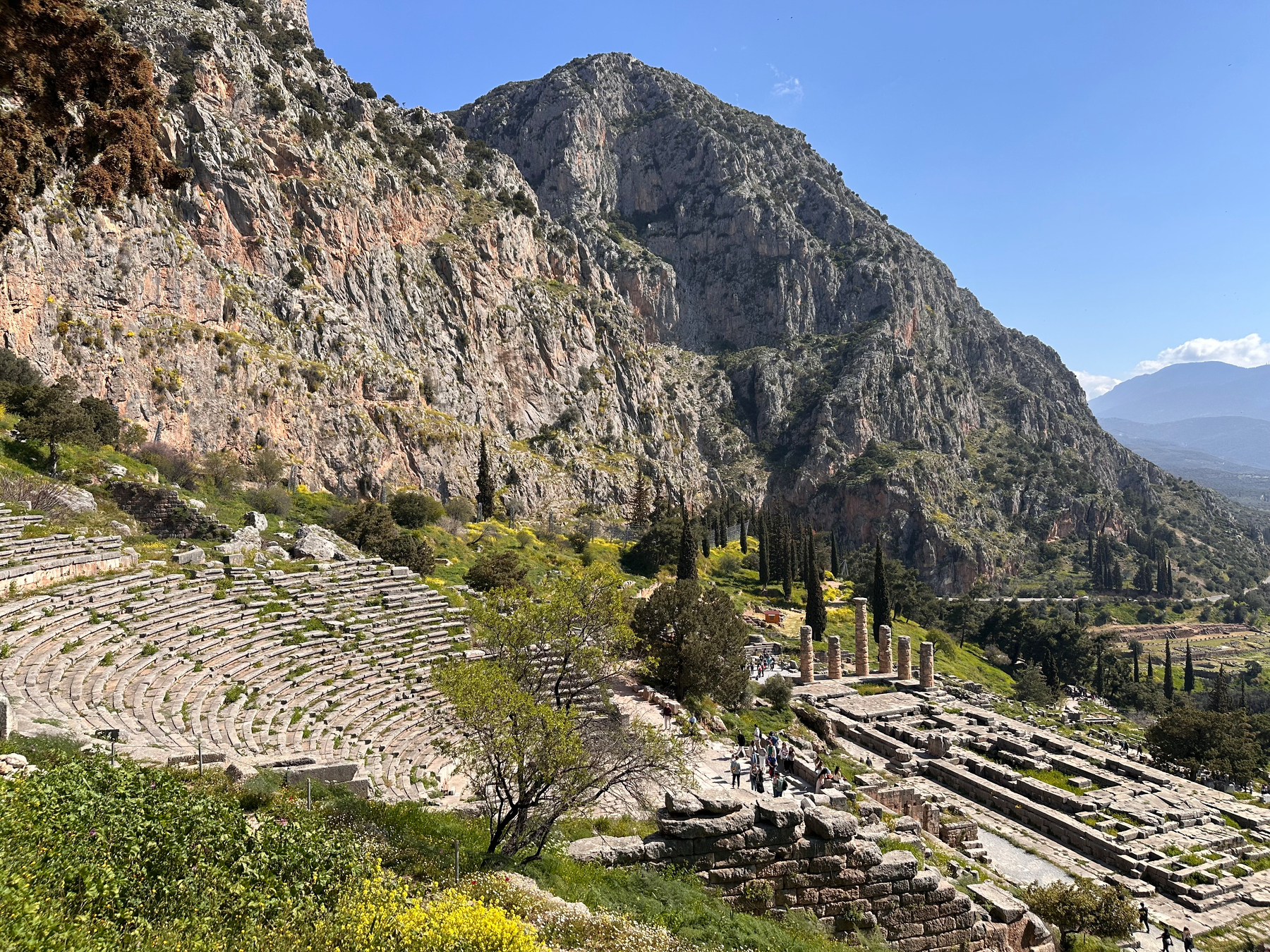 Ancient ruins, including a stone amphitheater, are set against a backdrop of rocky mountains and lush greenery.  Delphi, Greece. 