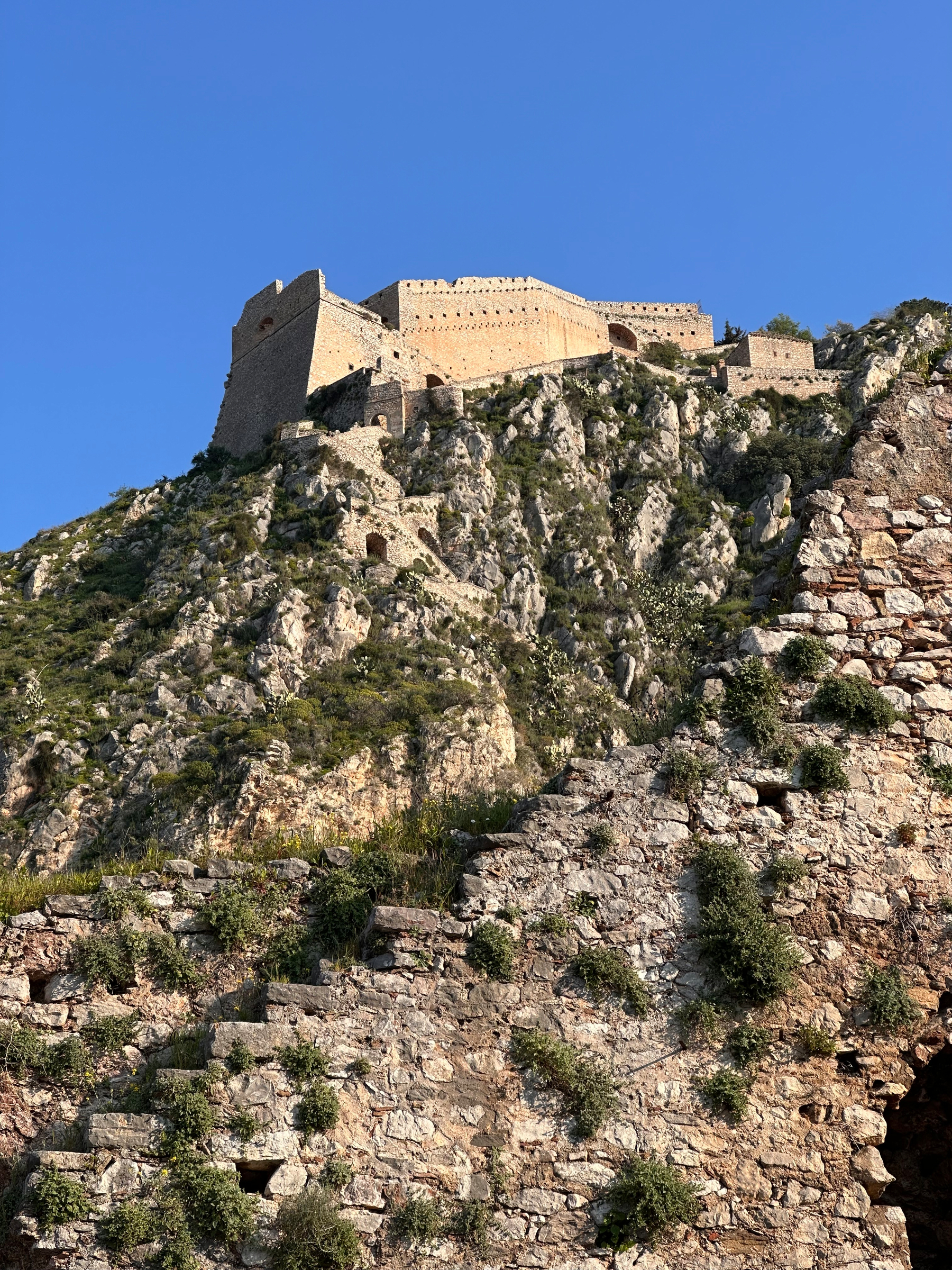 A majestic stone fortress sits atop a rugged, green hillside under a clear blue sky. Nauplion, Greece. Palamidi fortress. 