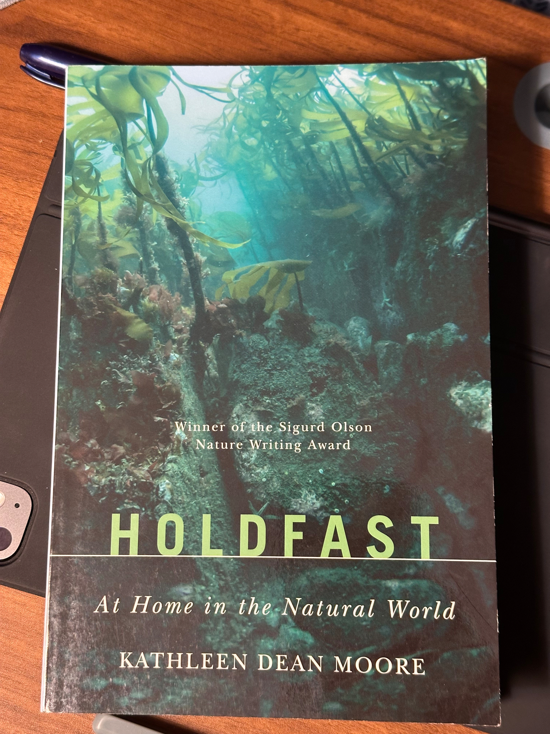 A book titled Holdfast: At Home in the Natural World by Kathleen Dean Moore, featuring an underwater scene with seaweed on its cover, rests on a wooden surface alongside a pen and an electronic device.