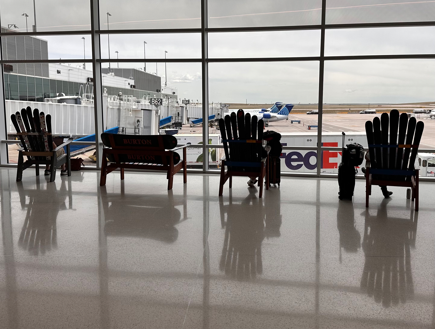 Several large Adirondack chairs made of skis and a bench made of snowboards are lined up in front of a large window overlooking airport gates and aircraft.