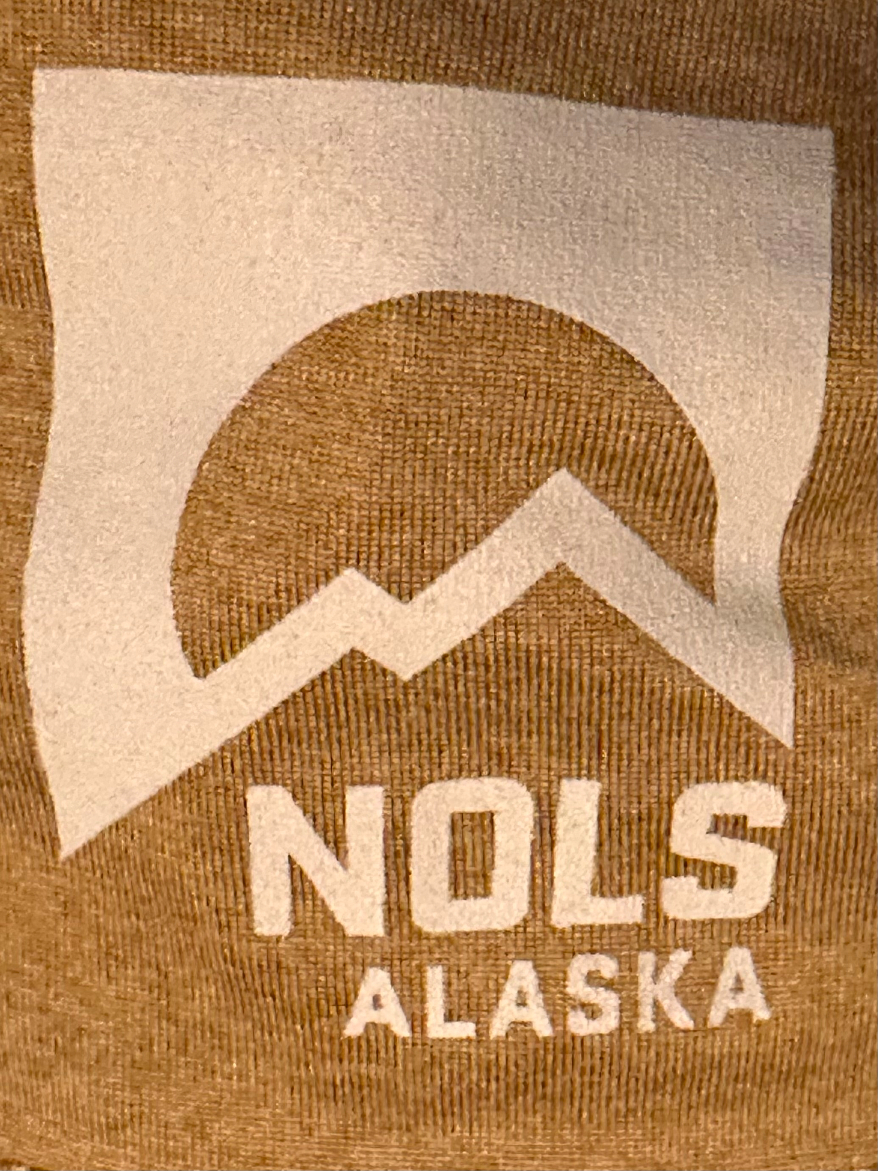 A logo featuring a stylized mountain and sun design with the text NOLS ALASKA beneath it is displayed on a brown textured fabric.