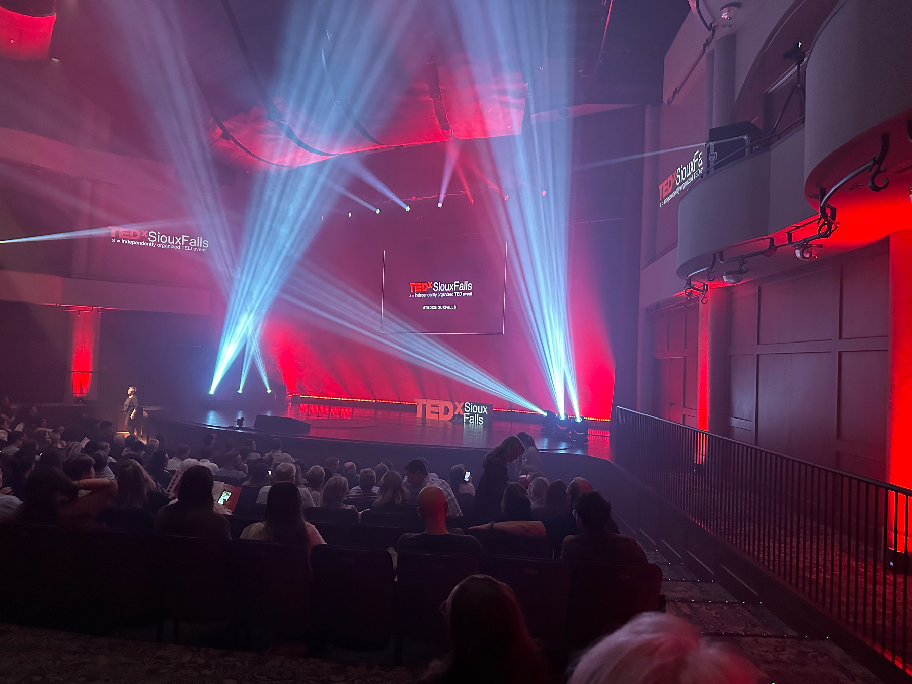 A TEDx event in Sioux Falls features a speaker on stage with an audience seated, surrounded by vibrant red and blue lighting.