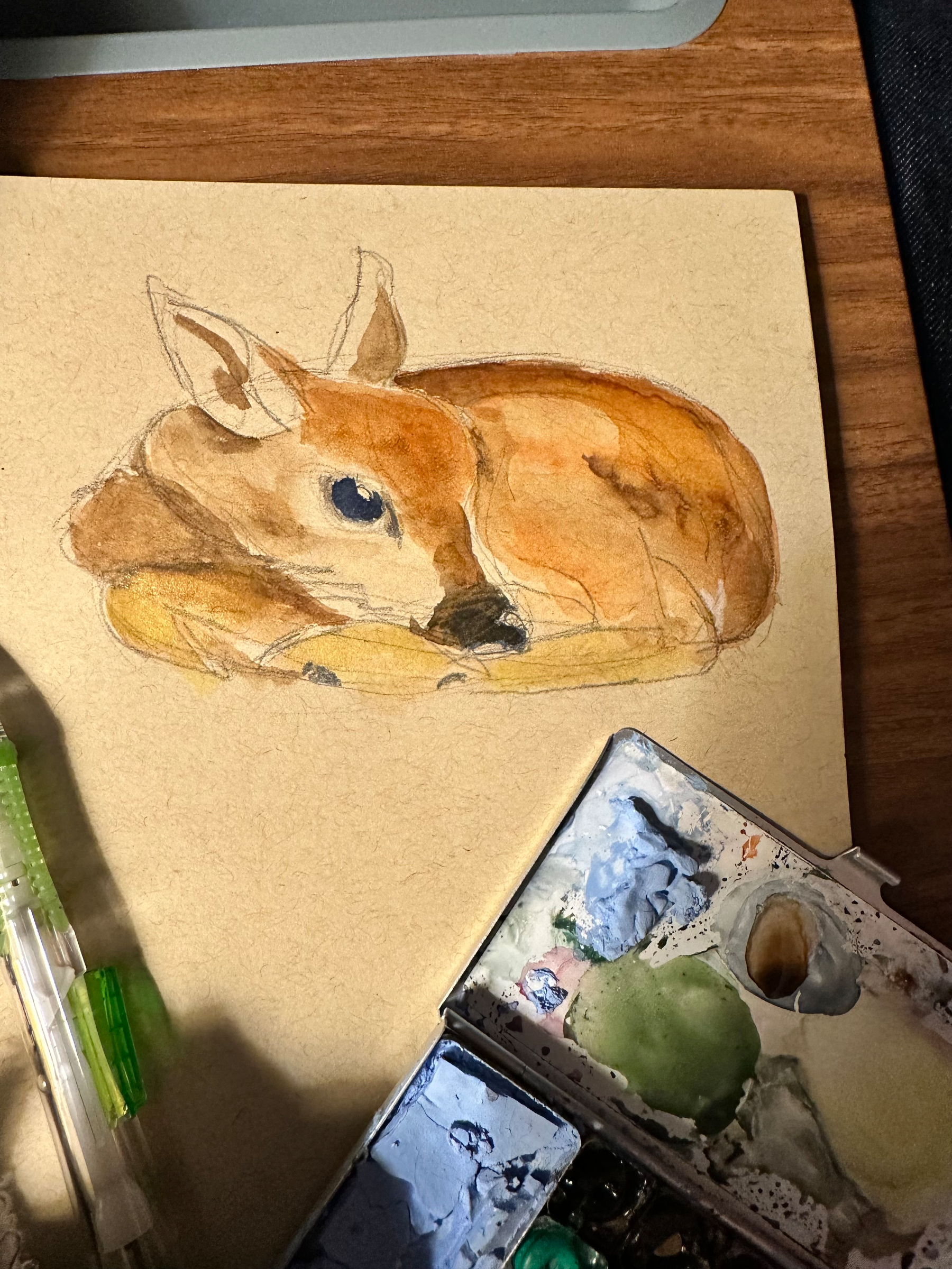 A watercolor painting of a resting deer is displayed on a piece of paper alongside a paint palette and a green pen.