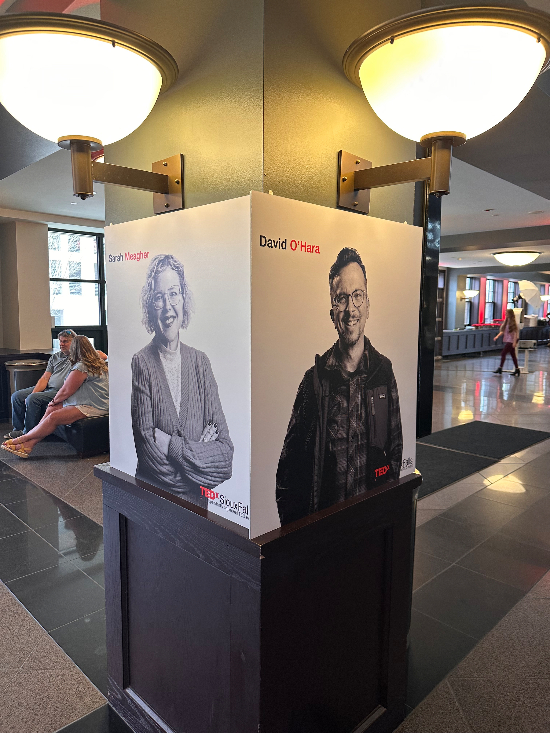Two poster displays showcasing individuals named Sarah Meagher and David O'Hara, both part of a TEDx Sioux Falls event, are positioned back-to-back in a public indoor area.