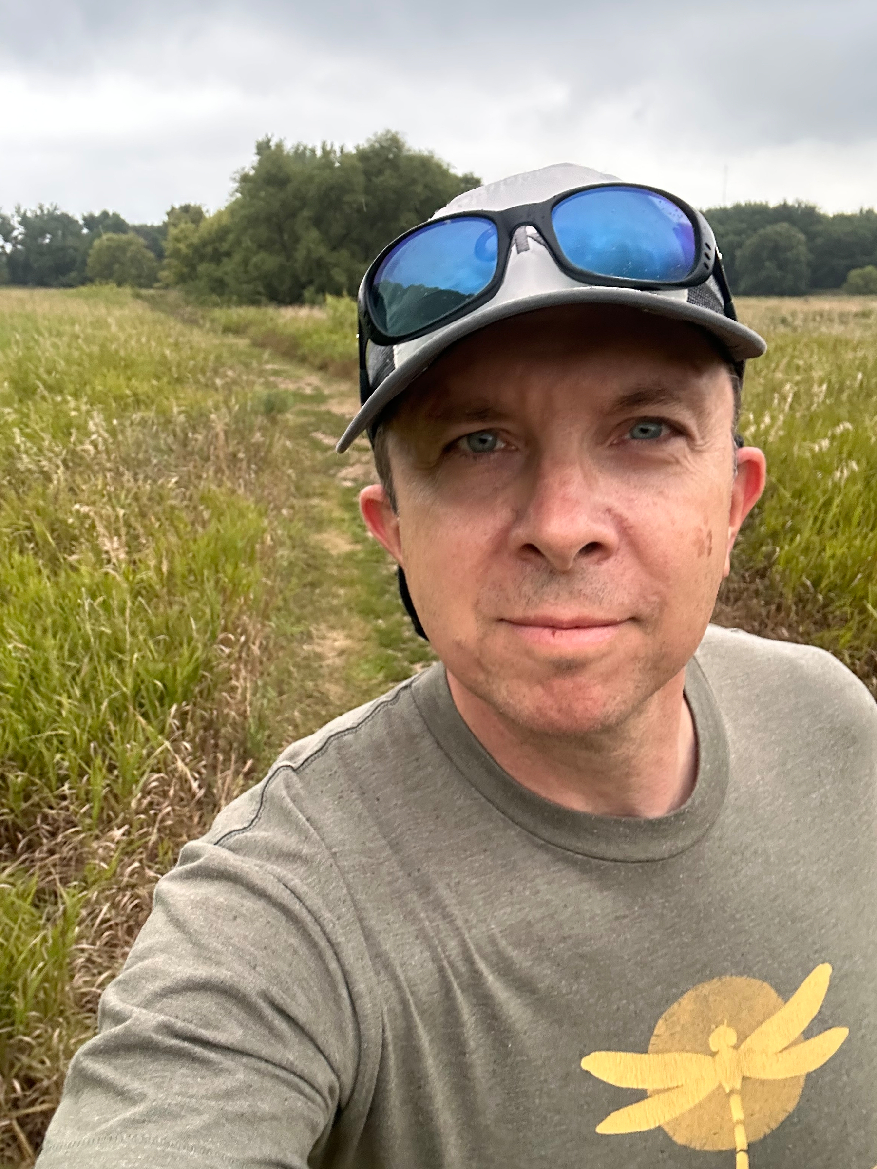 A man wearing a gray cap and a green shirt with a yellow dragonfly graphic is taking a selfie outdoors on a grassy trail with trees in the background under a cloudy sky.