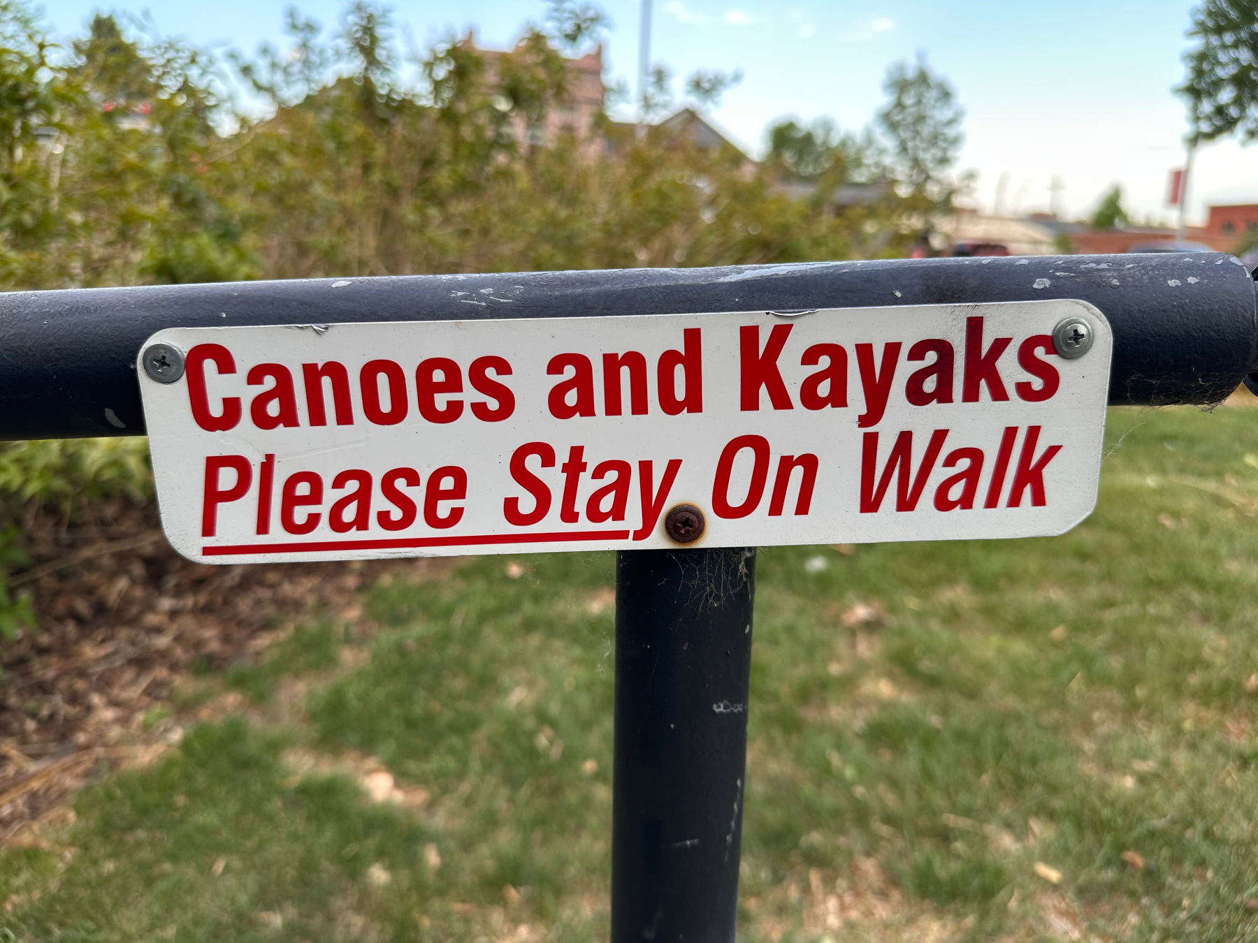Photo of a small sign screwed to a black metal handrail. The white metal sign has red lettering that reads “Canoes and Kayaks Please Stay On Walk” 