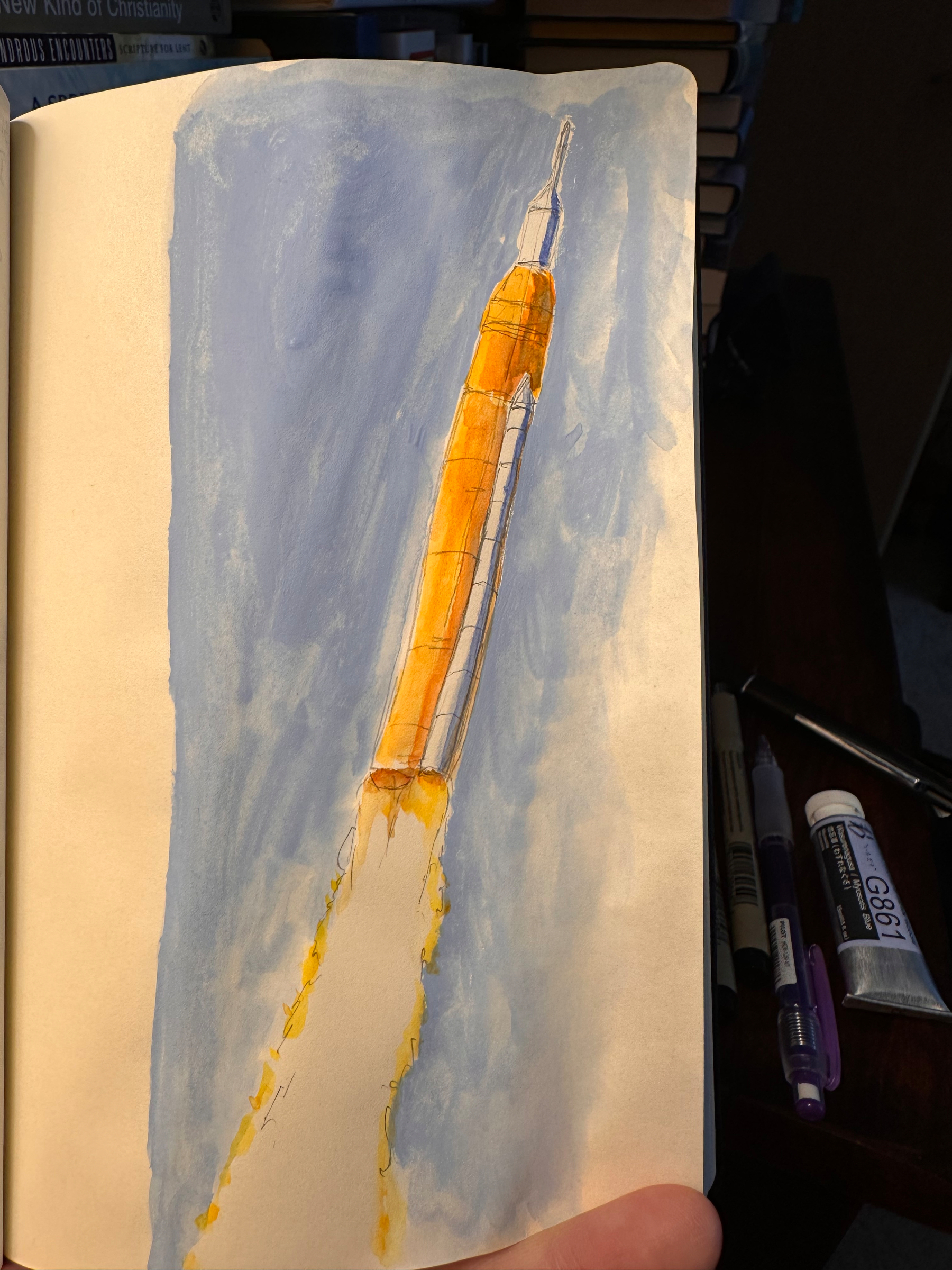 A hand-drawn illustration of a rocket launching, with blue smoke and a tall, orange body, against a blue background, is visible on a sketchbook page.