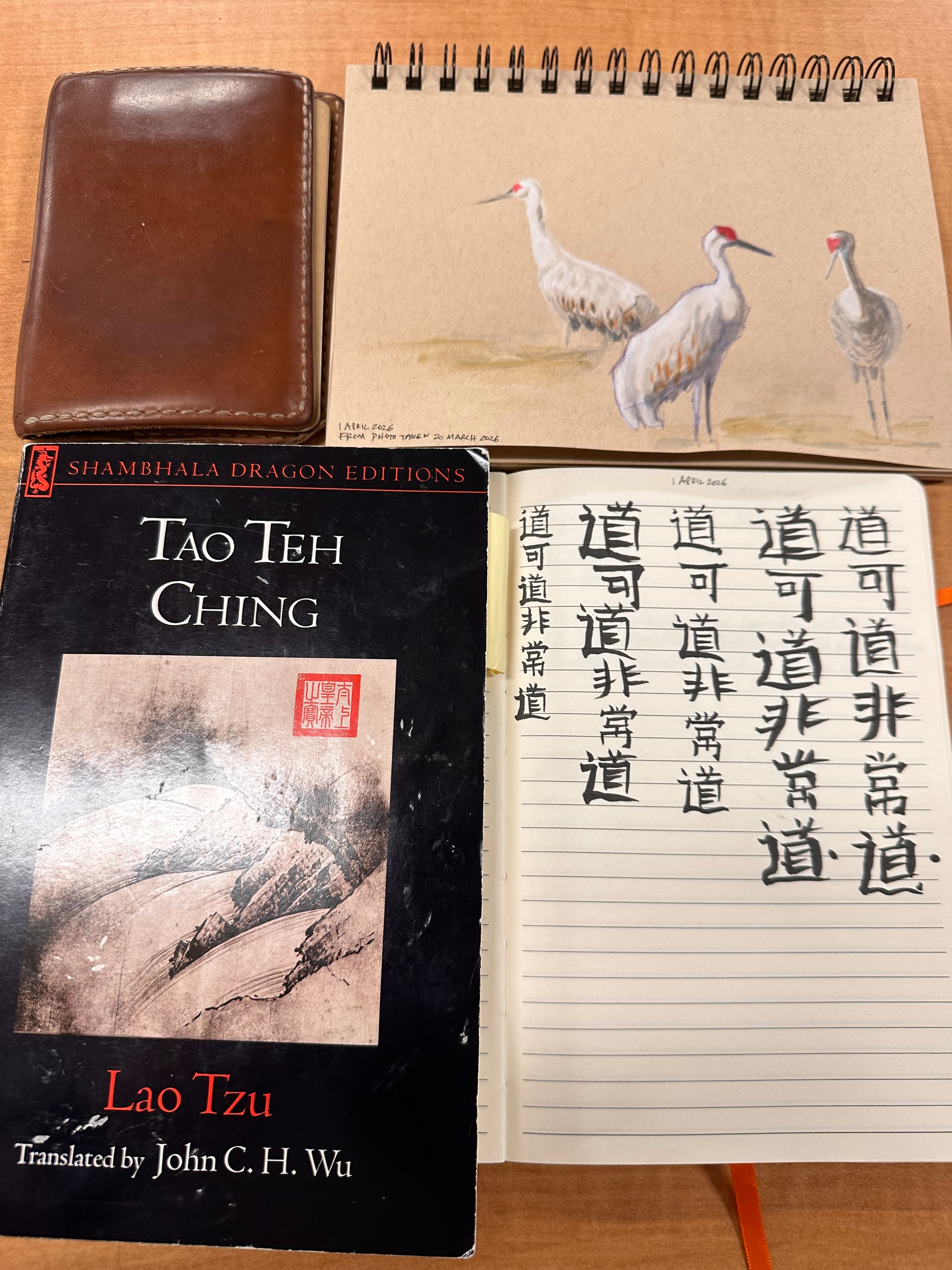 A notebook with handwriting in Chinese characters and a cover of Tao Teh Ching are placed beside a sketchpad featuring three cranes, along with a brown leather case.