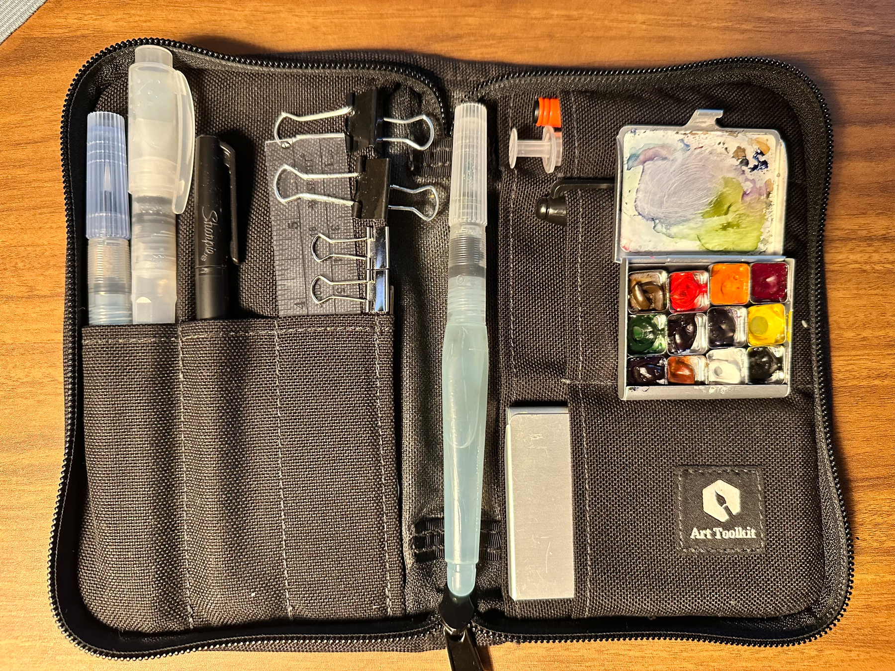 A black zippered art toolkit case contains various painting supplies including water brushes, ink pens, binder clips, a small watercolor palette, a mixing tray, and an eraser.
