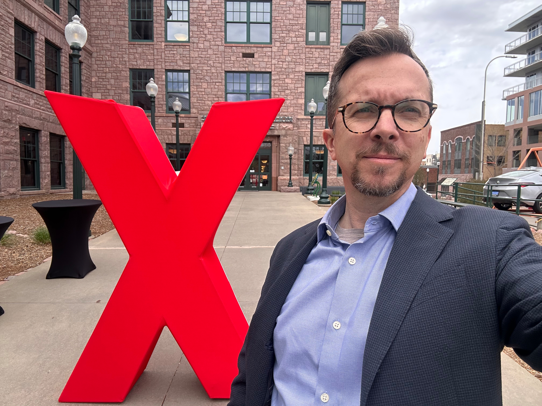 A person is taking a selfie in front of a large red X sculpture outside a brick building.