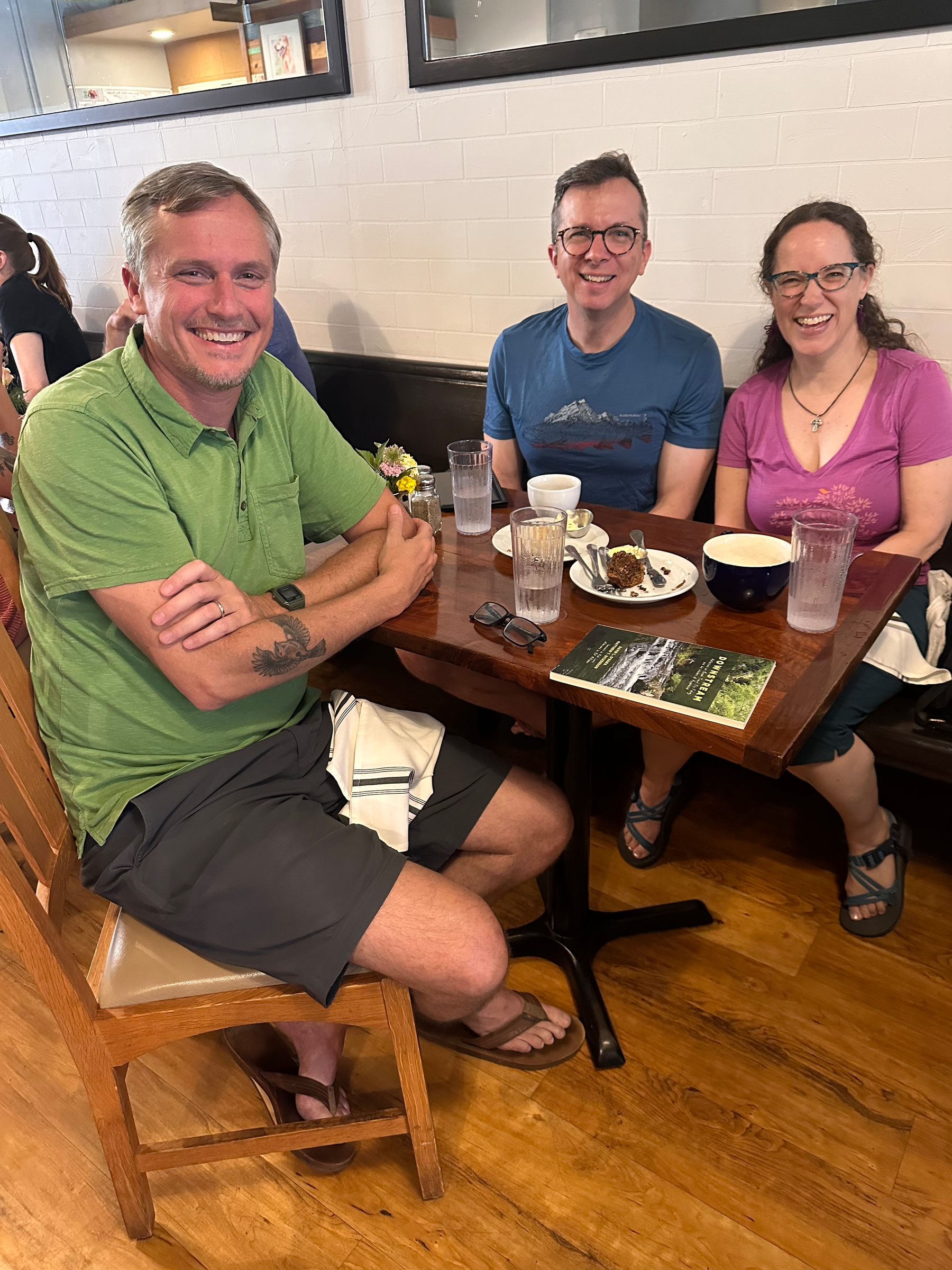 Three people are sitting around a wooden table in a casual dining setting, smiling at the camera with food and drinks in front of them.