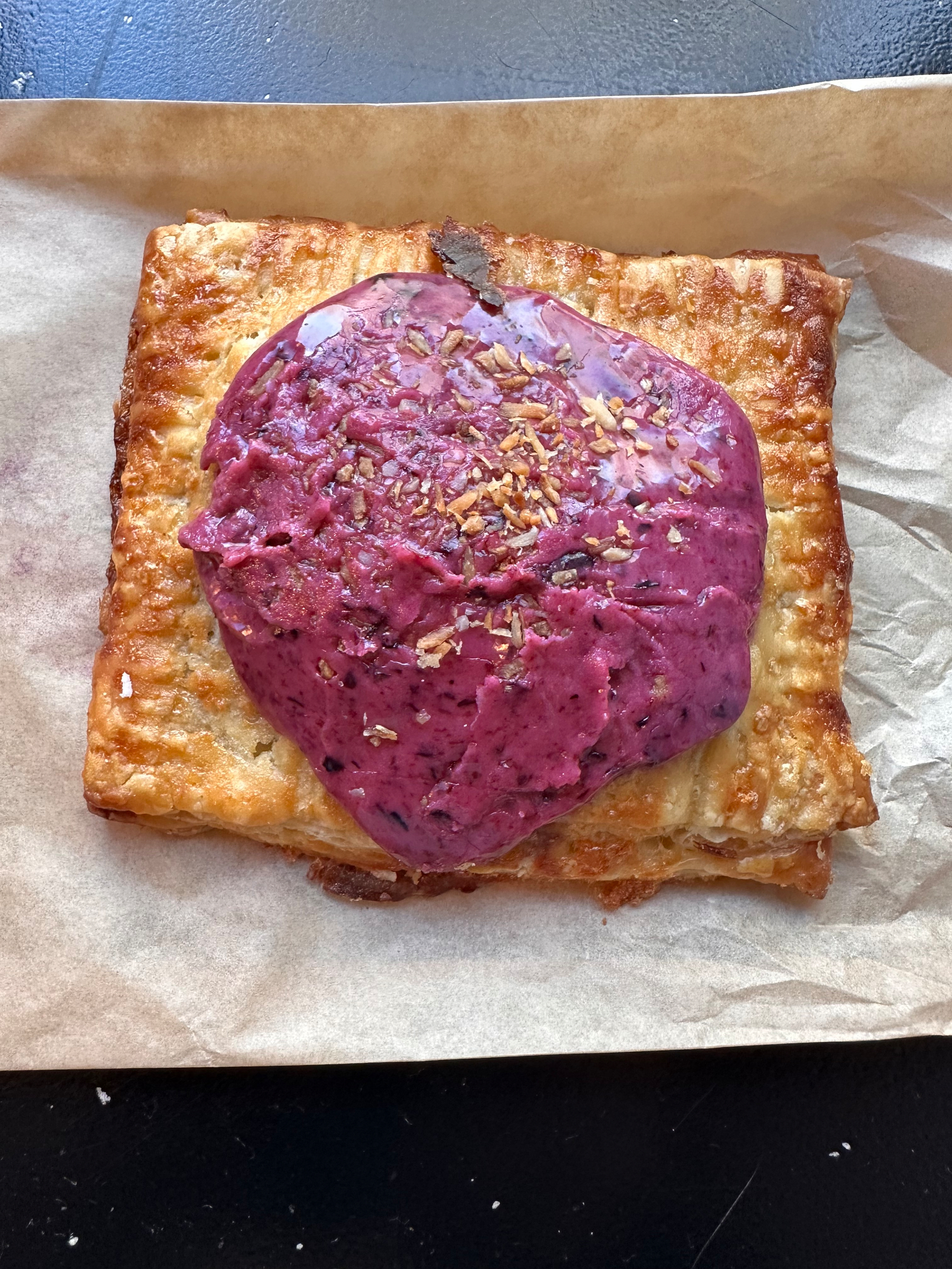 A toasted sandwich topped with a dollop of purple spread and sprinkled with crushed nuts sits on a piece of parchment paper.