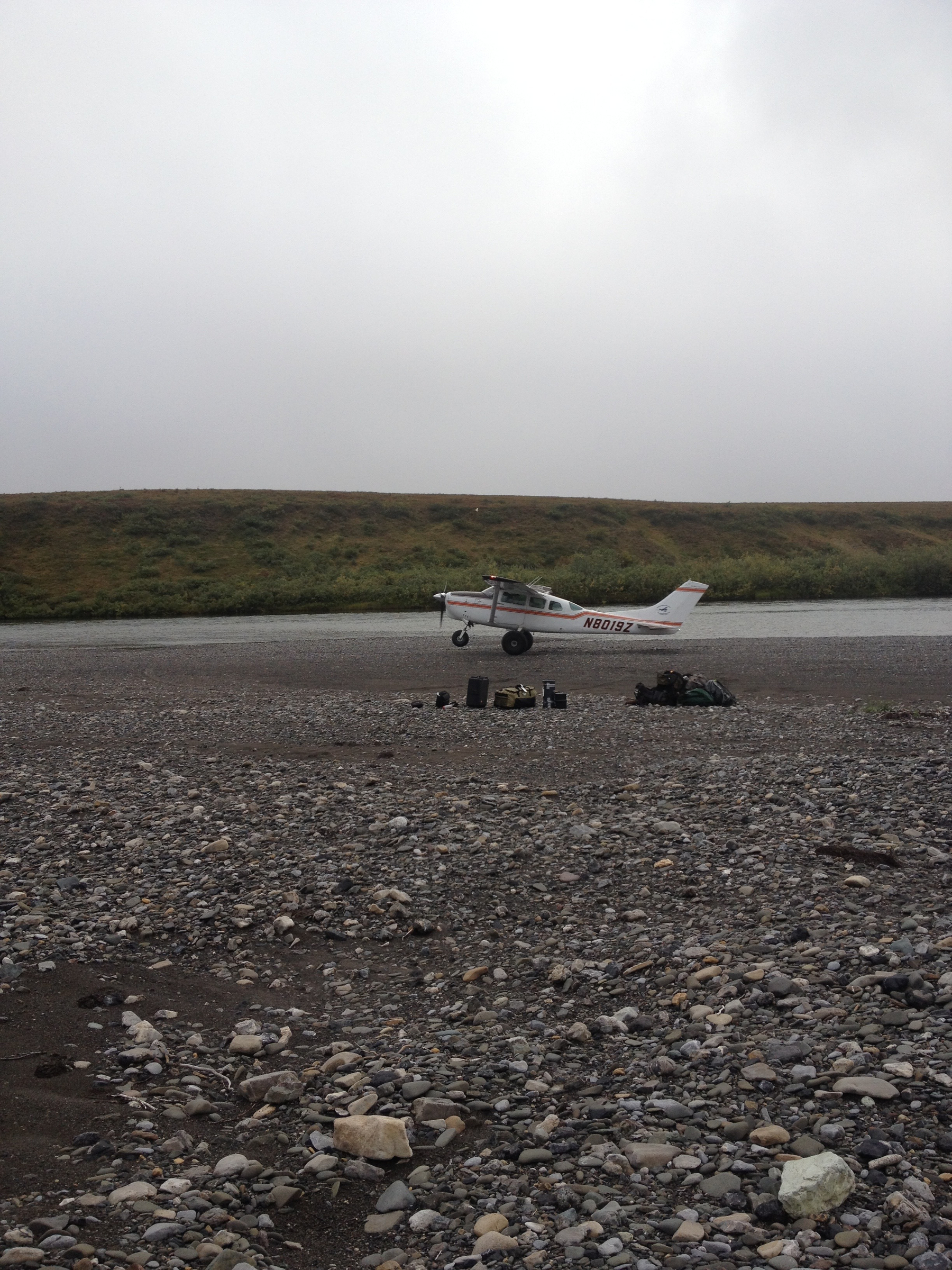A small single-engine airplane is landing or taking off on a riverbed with rocky terrain and some equipment or supplies placed nearby.