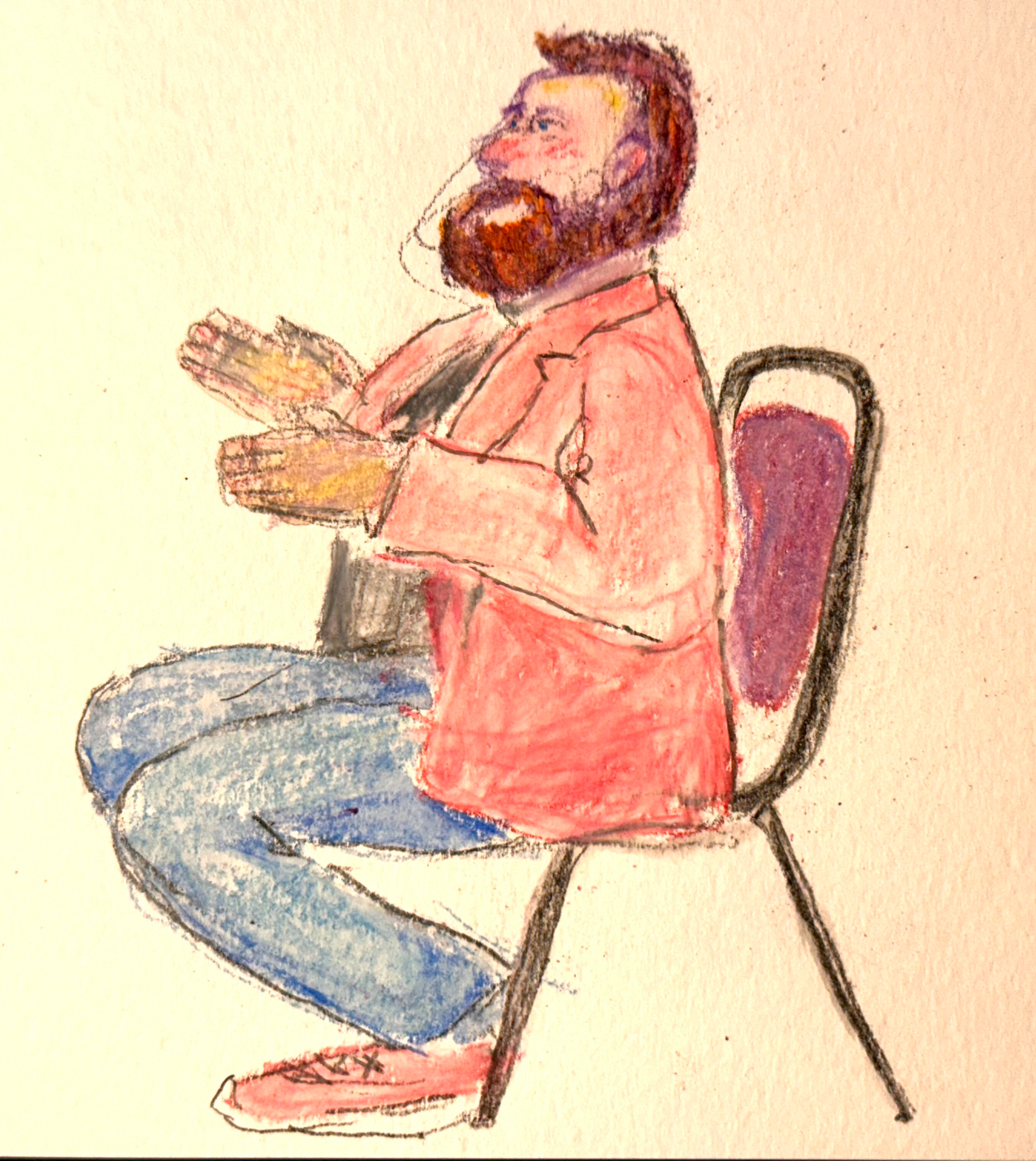 A bearded man wearing a red jacket and blue jeans is sitting on a chair and clapping his hands.