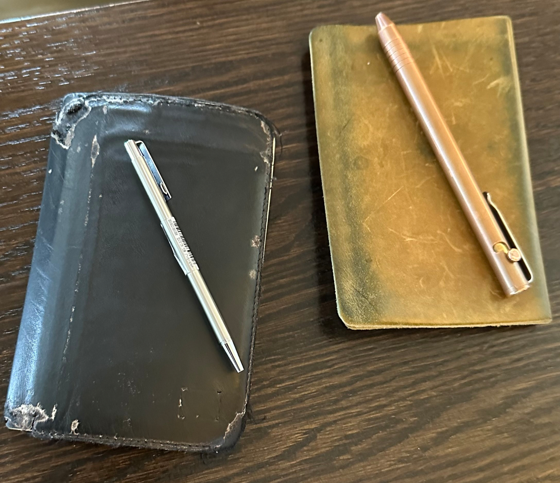 Two notebooks, one black with a silver pen and one brownish with a bronze pen, are placed on a wooden surface.