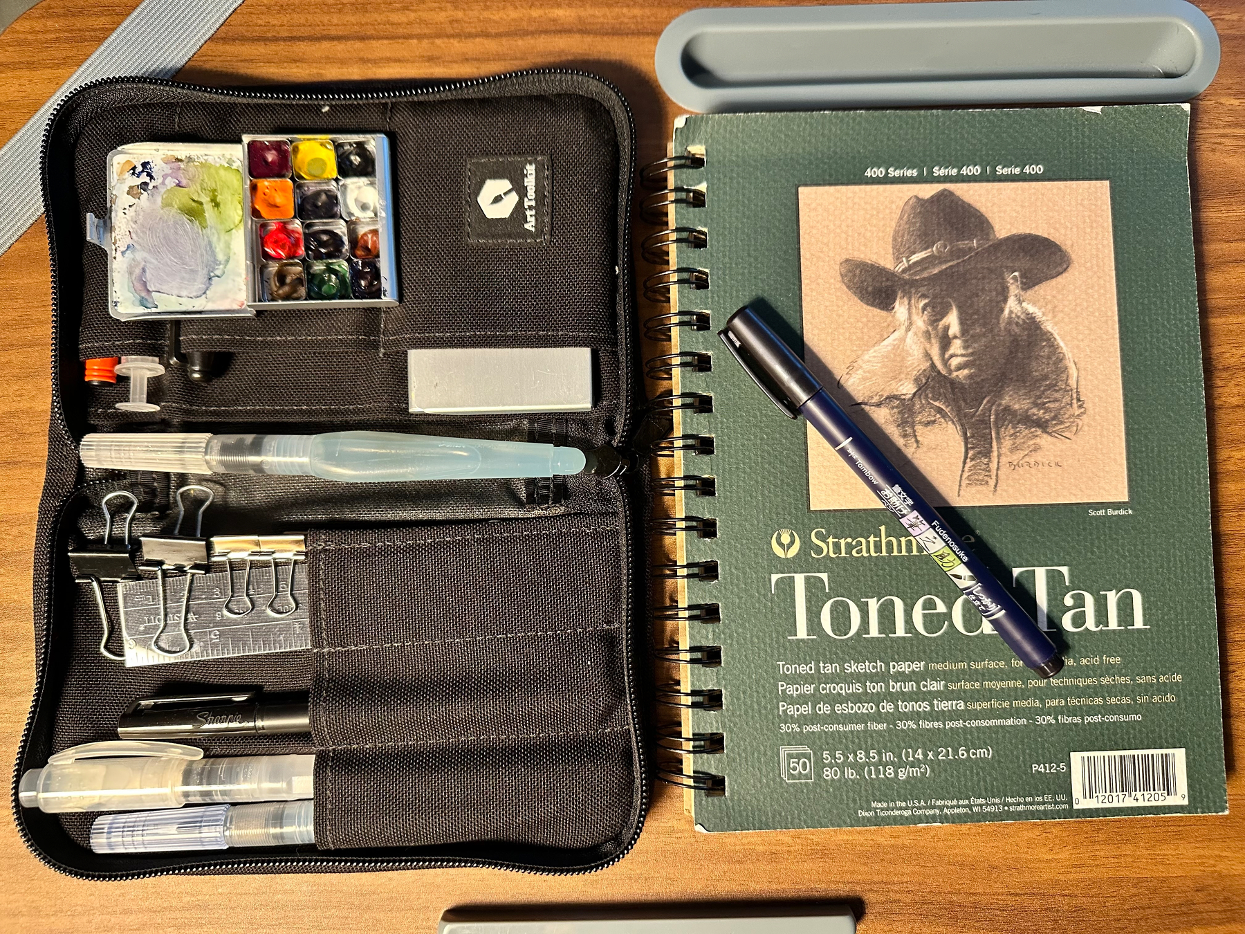 A watercolor paint set with brushes and clips is open next to a toned tan sketch pad with a pen resting on top of it.