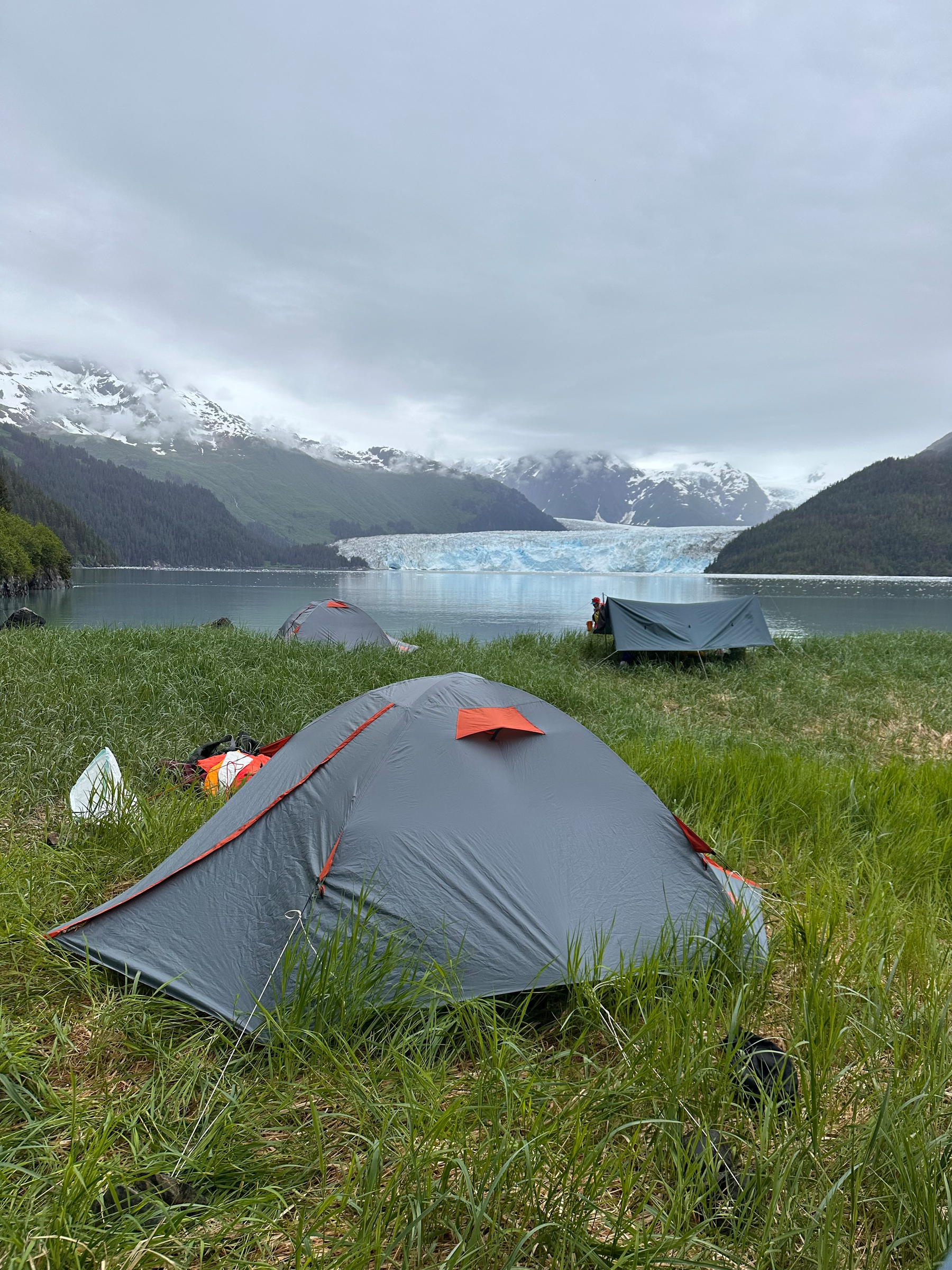 Several tents are set up on grassy terrain near a lake with a glacier and snow-capped mountains in the background under a cloudy sky.
