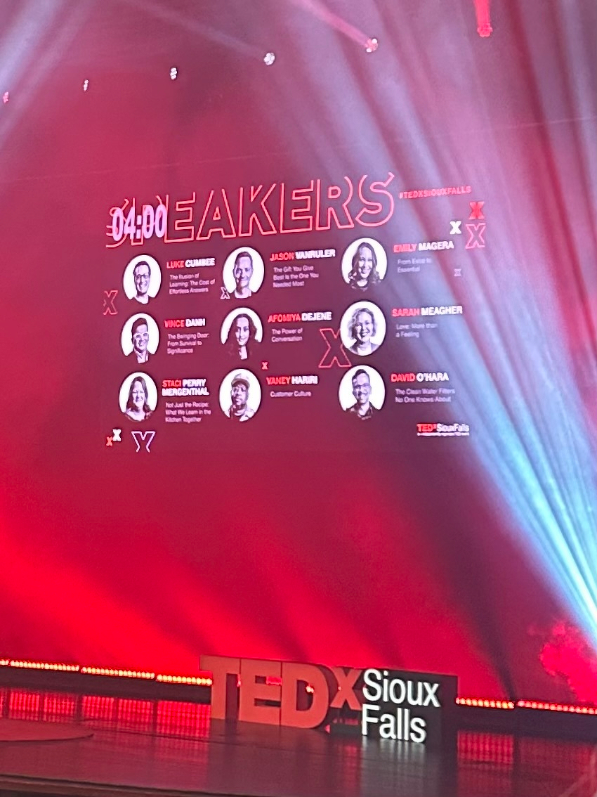 A stage is set for a TEDxSiouxFalls event, featuring a screen displaying information about speakers with red lighting.
