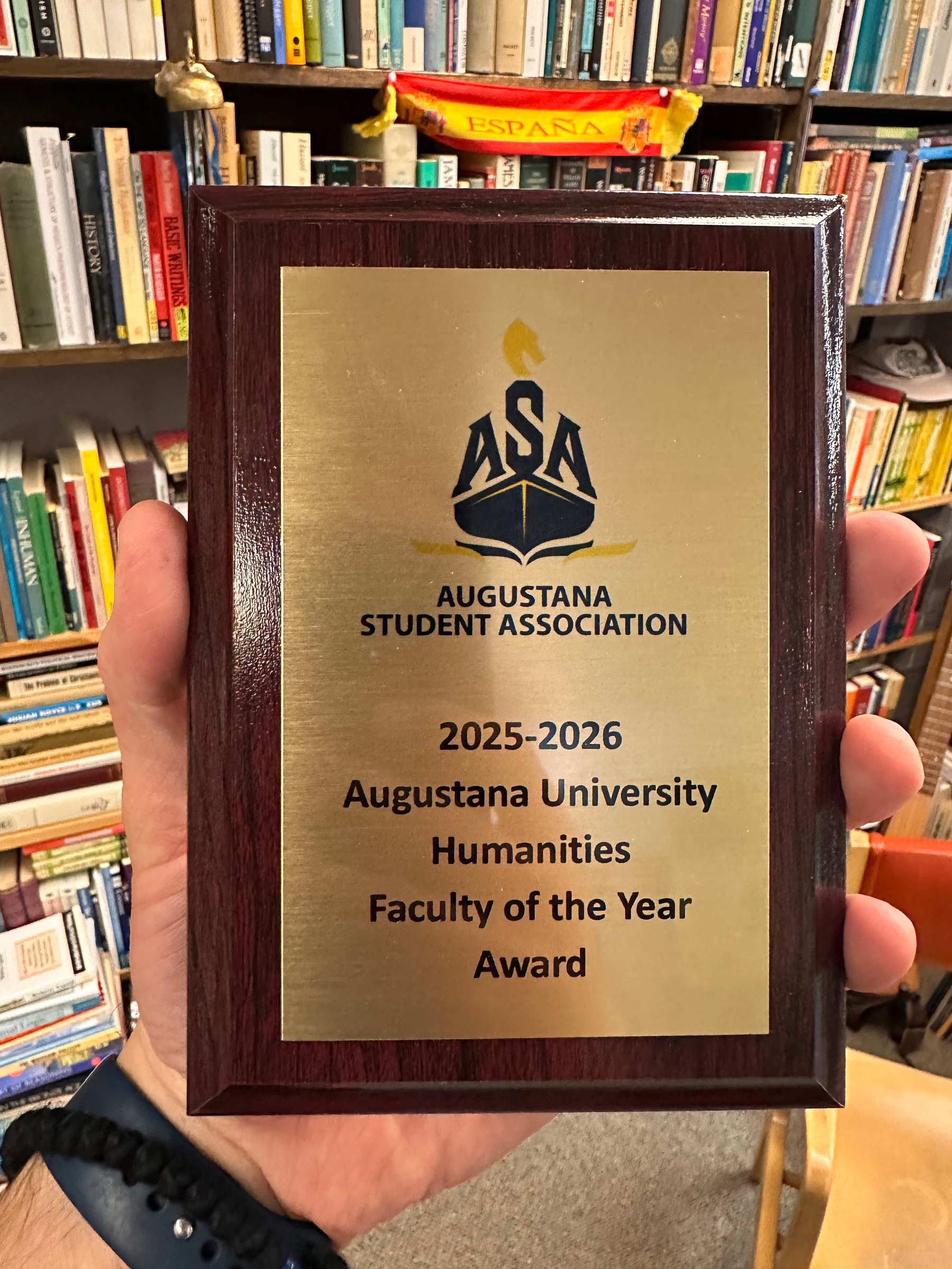 A hand holds a plaque awarded by the Augustana Student Association for the 2025-2026 Faculty of the Year in Humanities, positioned in front of a bookshelf.