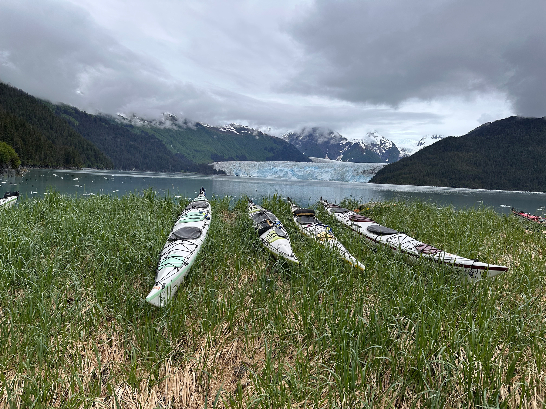 Several kayaks are resting on grassy terrain near a calm body of water with a glacier and snow-capped mountains in the background under a cloudy sky.