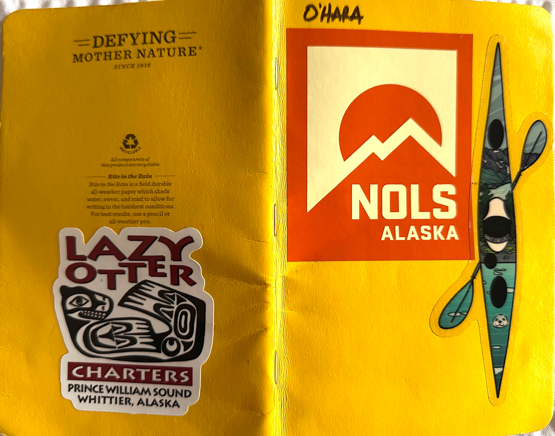 A bright yellow Rite-in-the-Rain field notebook splayed open so front and back covers are visible. On the front in bold black sharpie is the name “O’HARA” accompanied by a NOLS Alaska sticker and a sticker of a sea kayak and paddle that are both artistically decorated. The back cover has a sticker from Lazy Otter Charters, Prince William Sound, Whittier, Alaska. That sticker also has an indigenous-inspired (or indigenous-created?) line-art print of a sea otter.