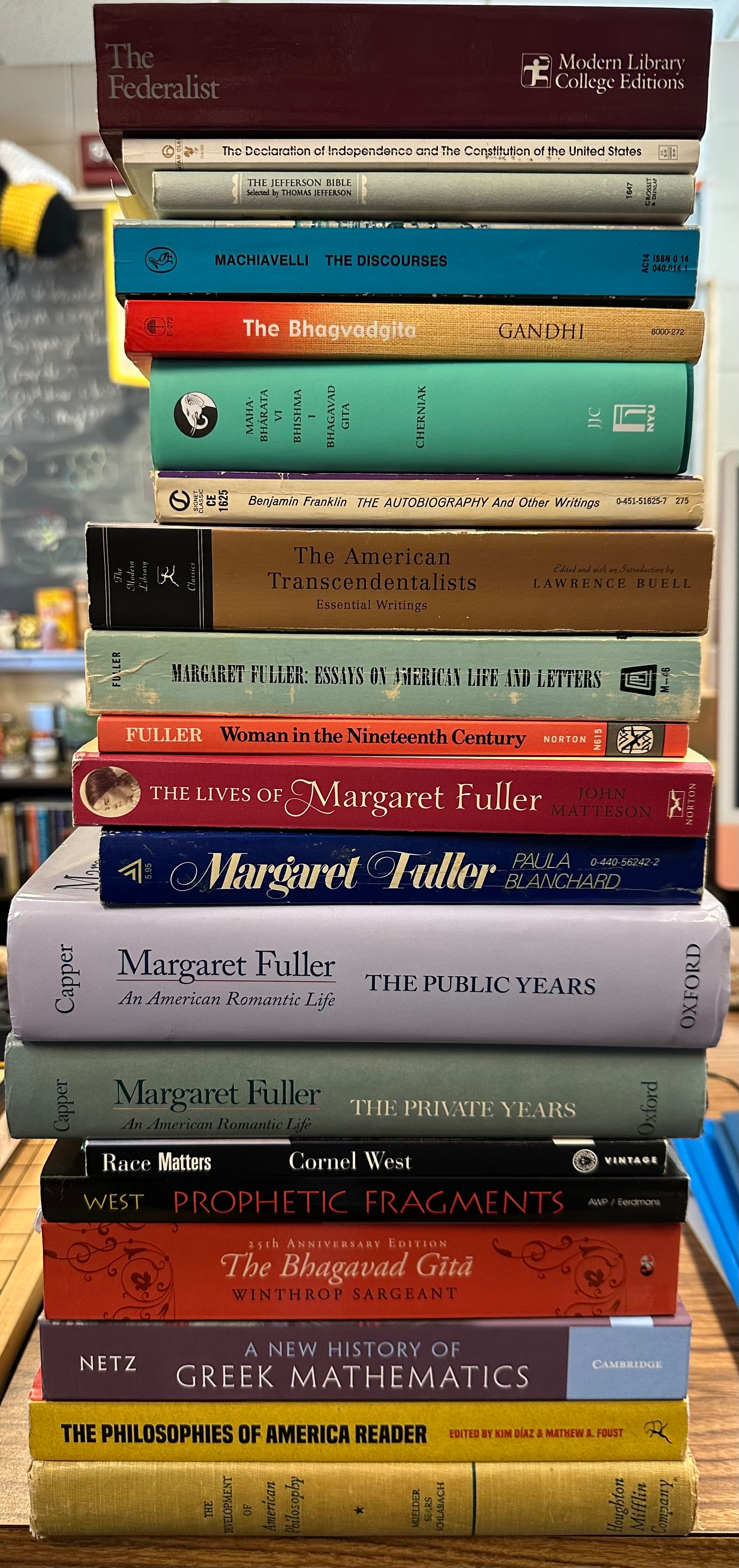 A stack of various books, including works by authors like Machiavelli and Margaret Fuller, and including several editions of the Bhagavad Gita, a work on the history of Greek mathematics, the U.S. Constitution, the Federalist Papers, and collections of works in Transcendentalism and American philosophies  is neatly arranged.
