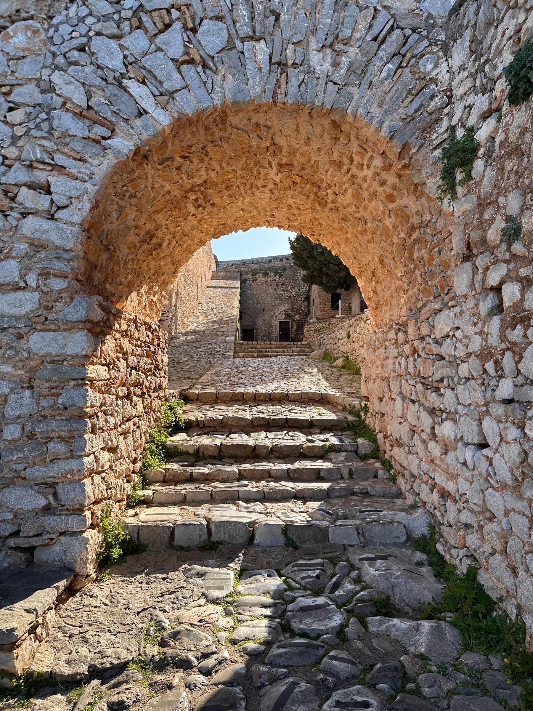 An ancient stone archway leads to a sunlit pathway with steps surrounded by rustic walls.