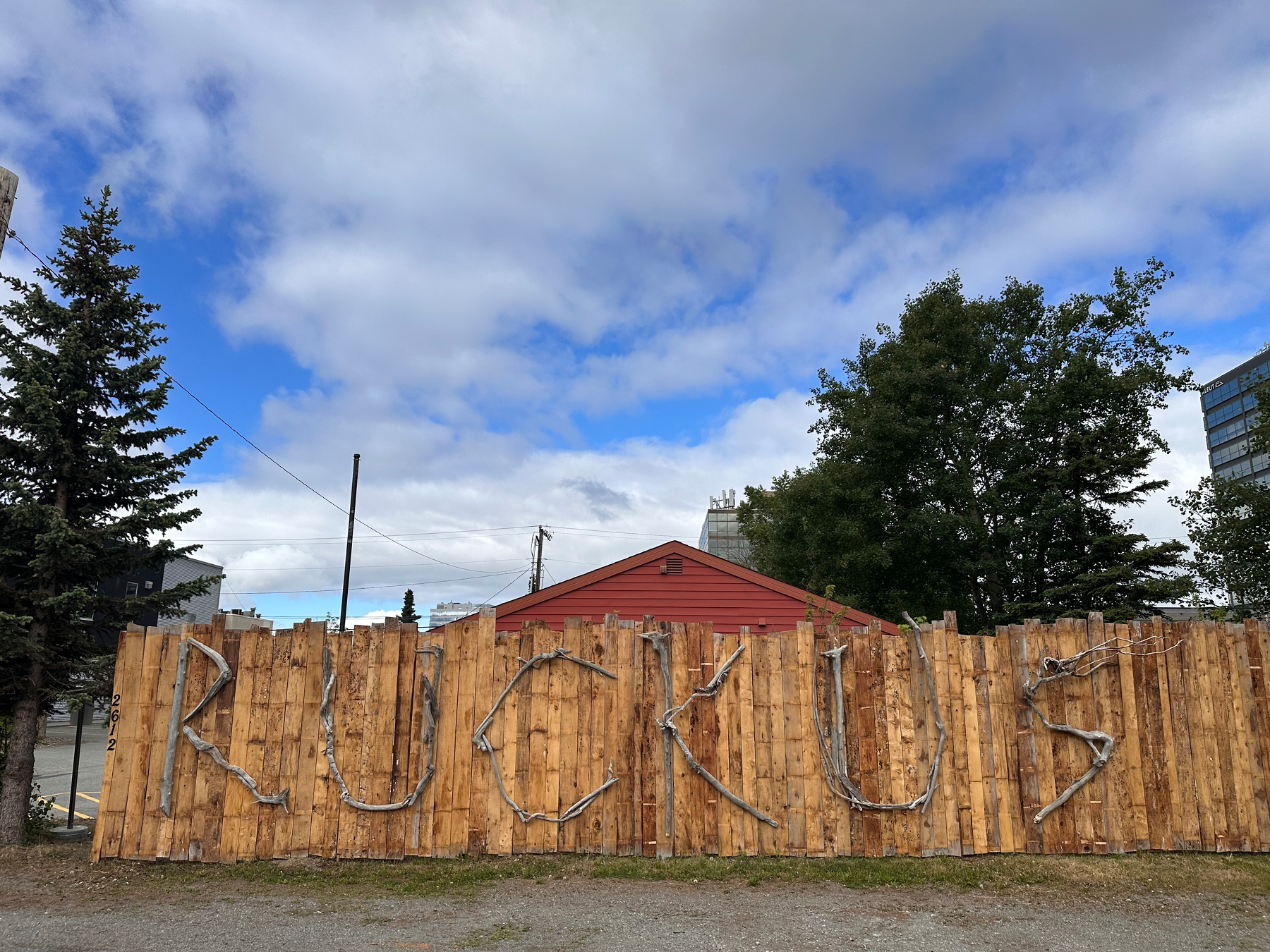 A wooden fence displays the word RUCKUS spelled out using sticks or branches against a background of trees, a red building, and a partly cloudy sky.