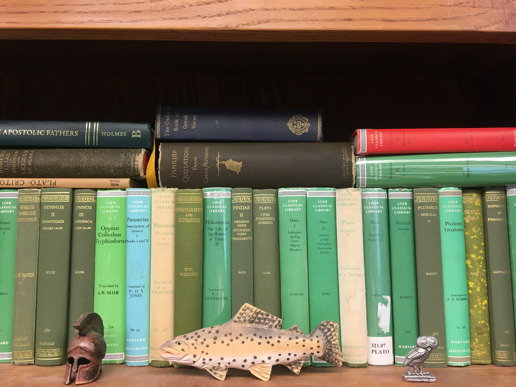 A wooden shelf filled with a variety of old hardcover books and a wooden fish sculpture along with a small metal figurine of an owl.