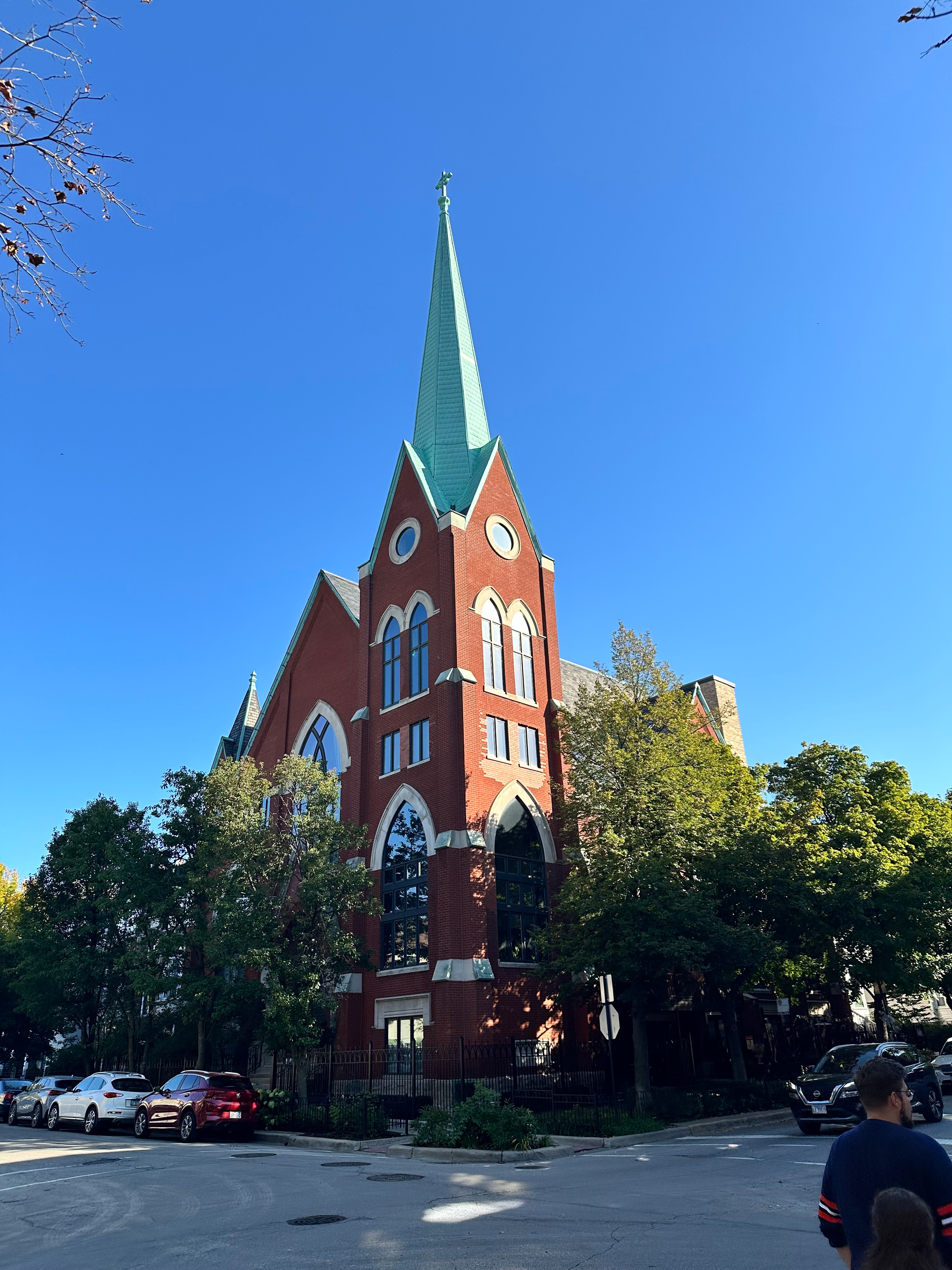 A tall red brick church with a green steeple stands prominently at a street corner surrounded by trees and parked cars under a clear blue sky.