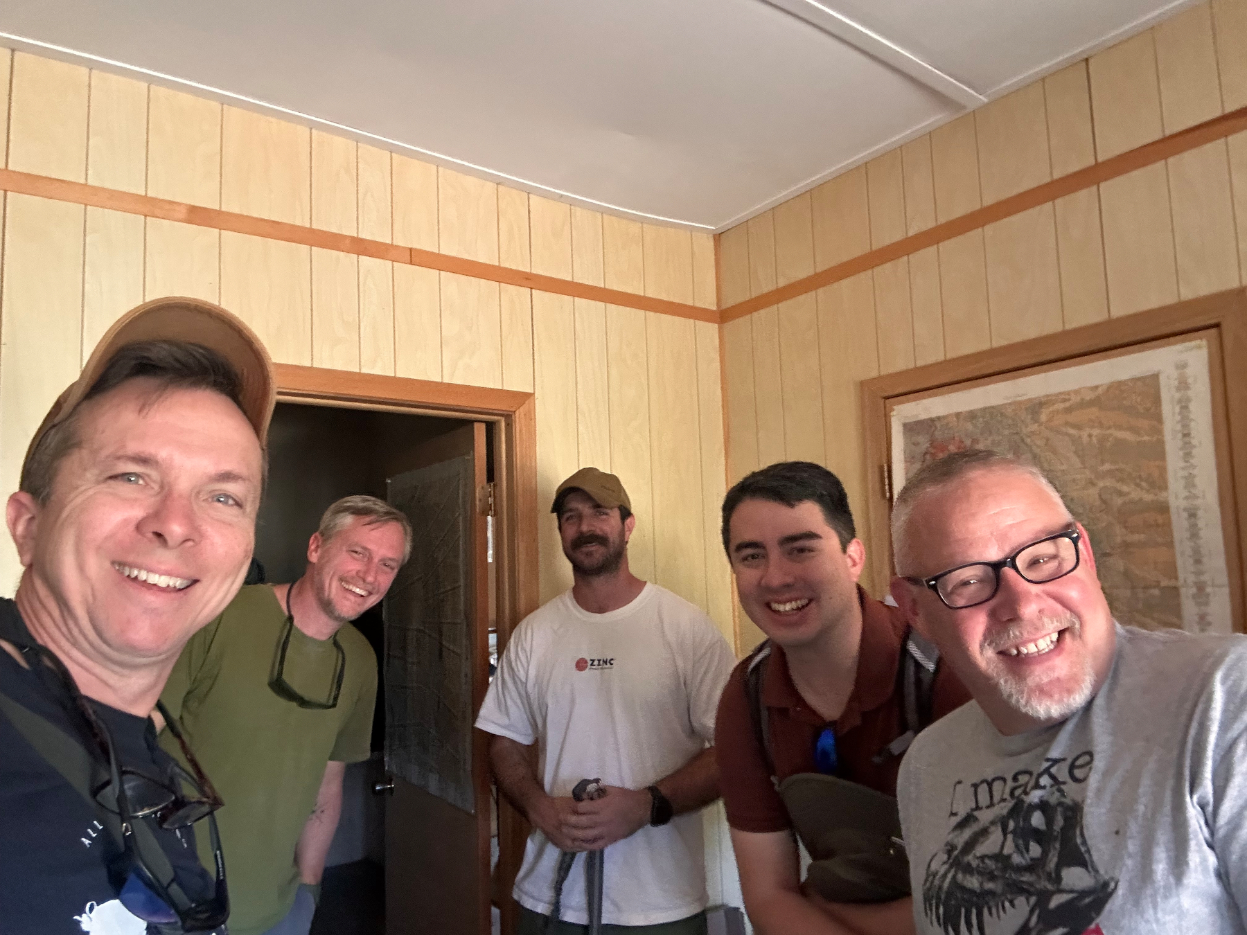 Five men are standing close together in a room with wood-paneled walls, smiling at the camera. Behind them is a geological map of the Paha Sapa or Black Hills, South Dakota.