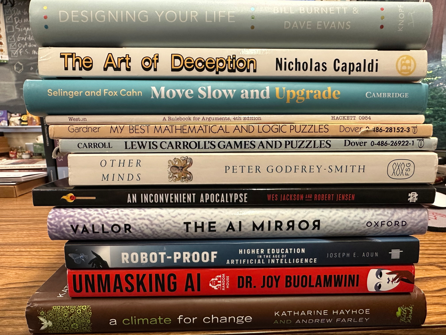 A stack of books on a wooden table covers topics ranging from AI and deception to puzzles and environmental issues.