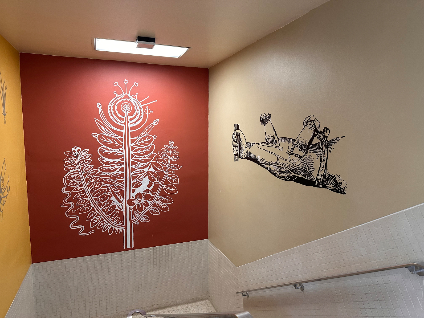 A staircase area features a wall mural with an intricate white botanical design on a red background and a hand-drawn illustration of a hand holding small anthropomorphic figures on a beige wall.