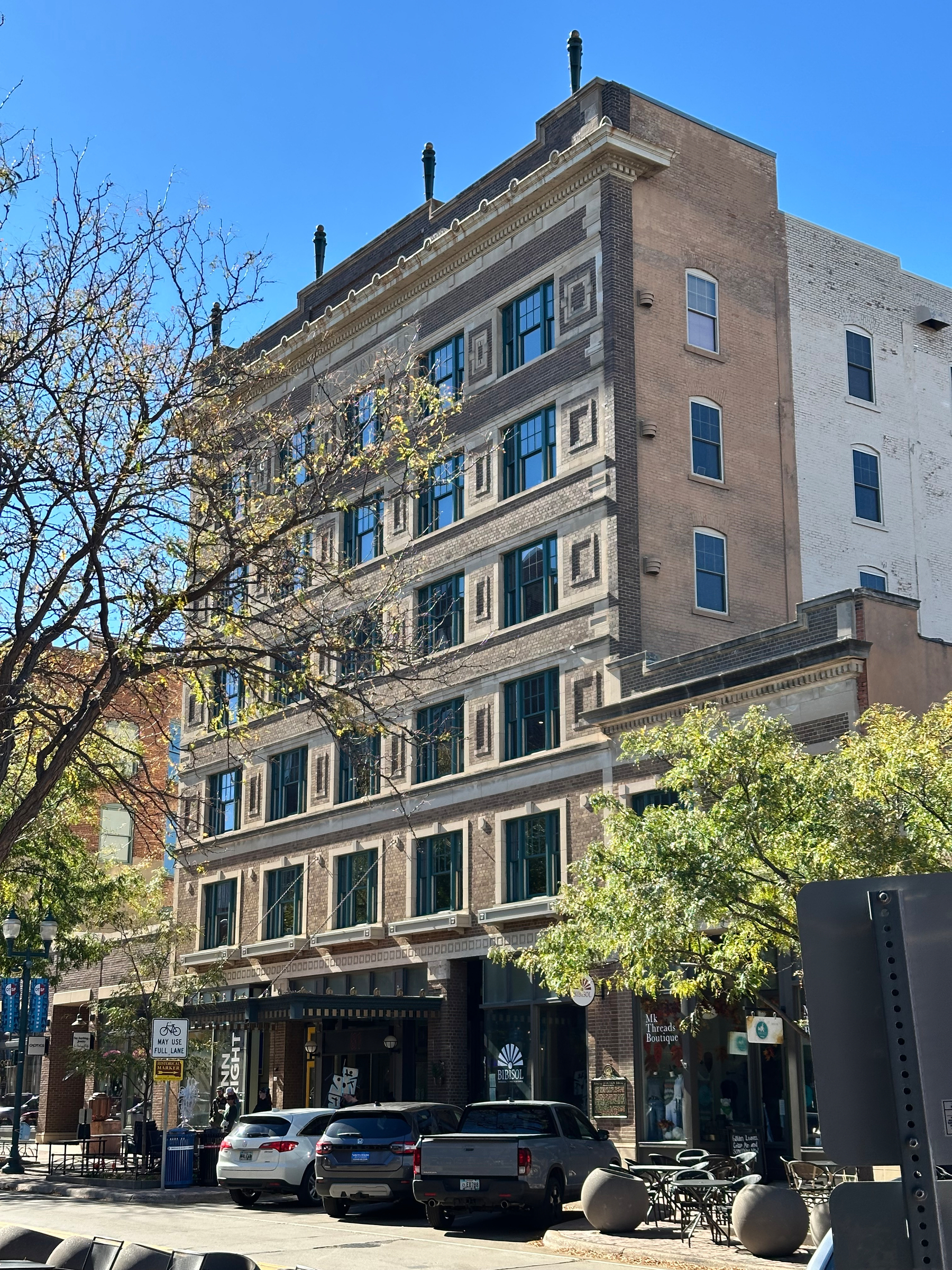 A historic multi-story brick building with decorative window frames stands on a sunny street lined with trees, parked cars, and outdoor seating.