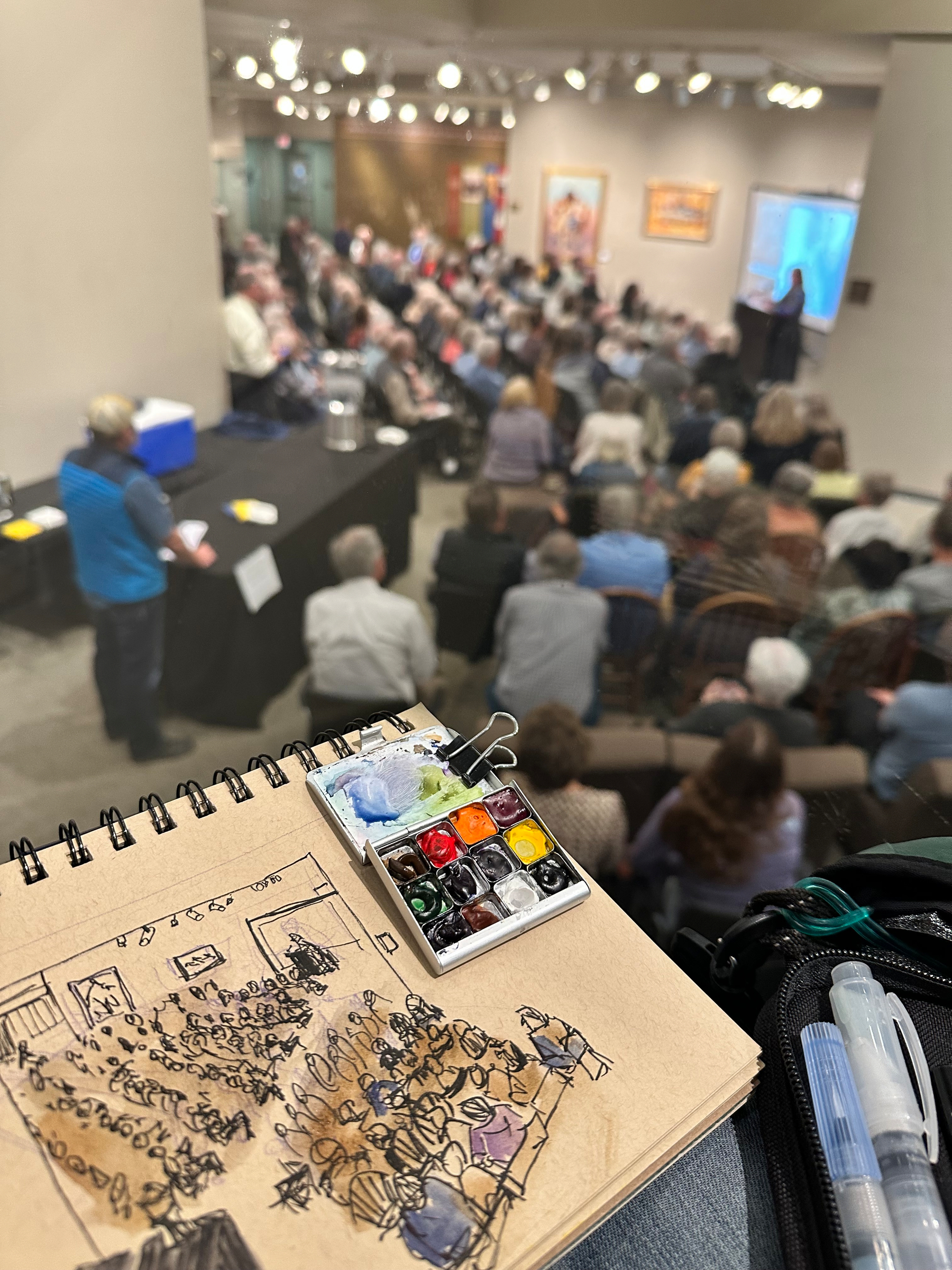 A person is sketching and painting a live scene of a crowded lecture or presentation from an elevated viewpoint.