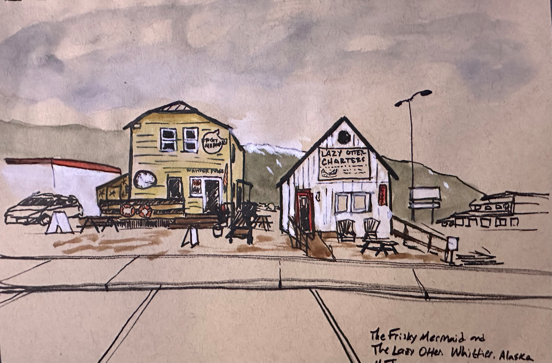 Two rustic buildings labeled The Frisky Mermaid and Lazy Otter Charters stand side by side in a small, quiet town with picnic tables and distant mountains in the background.